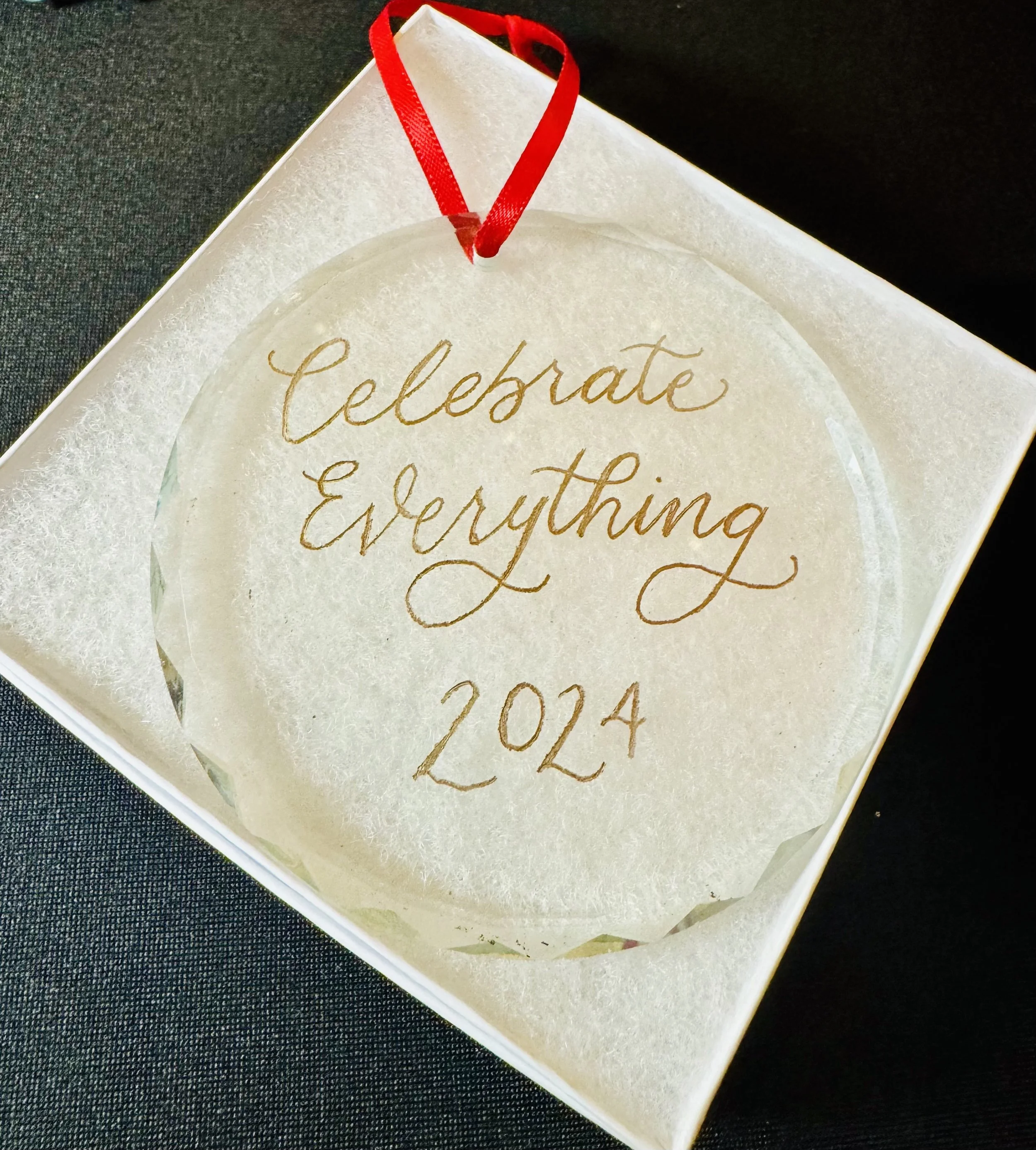 A decorative ornament with the inscription "Celebrate Everything 2024" in gold lettering, hanging from a red ribbon inside a white box with a padded interior.