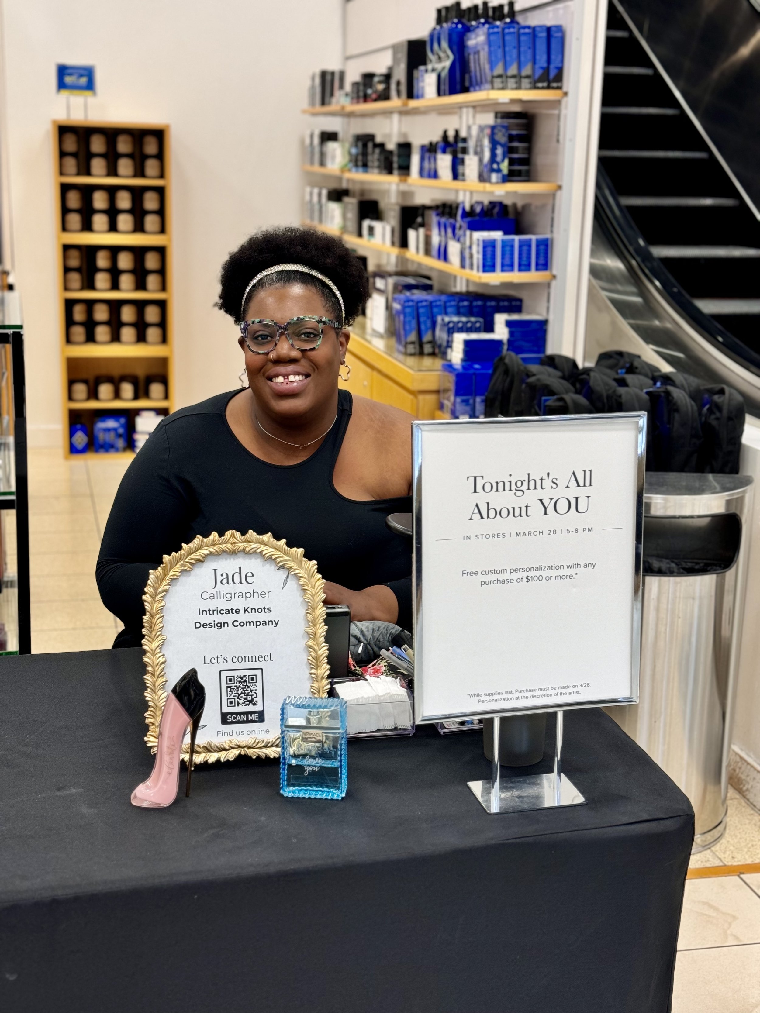 A smiling woman with glasses and an afro hairstyle sitting at a table with promotional signs and items. She is in a store or mall with shelves holding blue and black products behind her, near an escalator.