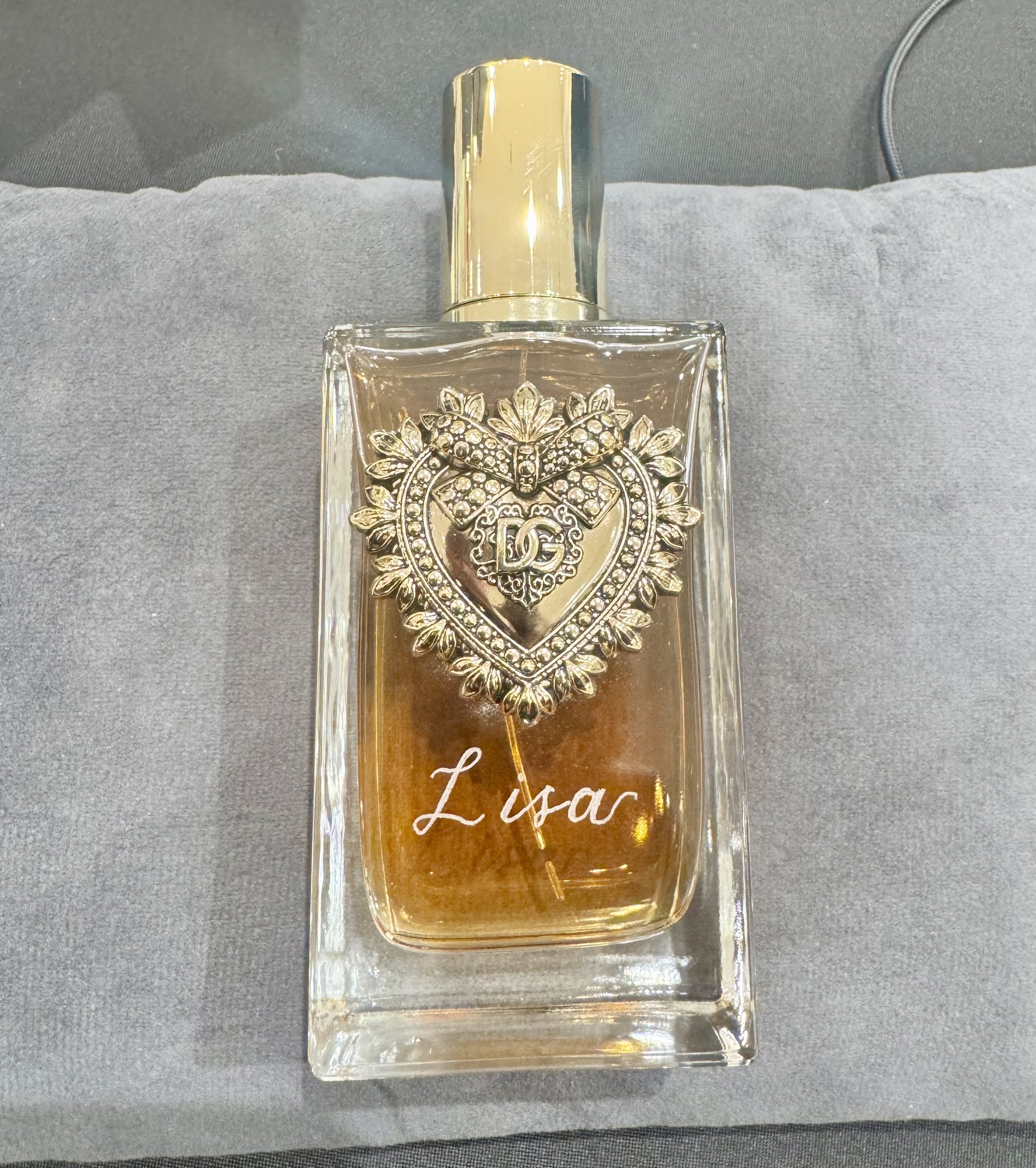 A clear glass perfume bottle with a silver decorative heart and bow embellishment on the front, labeled 'Lisar' in cursive writing, resting on a soft gray surface.