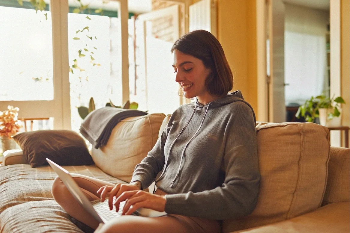 Eine Frau sitzt lächelnd auf einem Sofa und tippt in einen Laptop