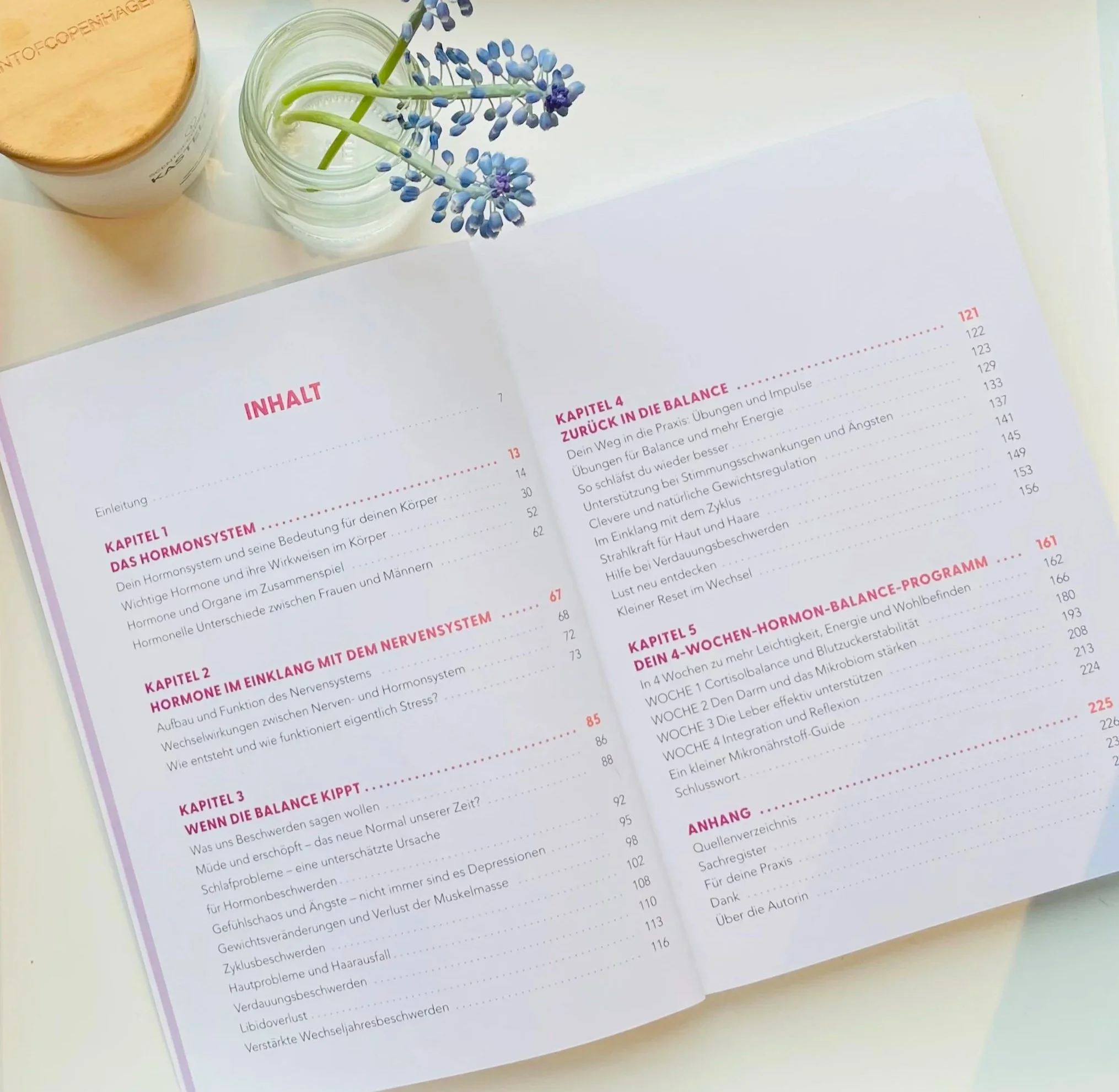 Das Buch "Neustart für die Hormone" von Astrid Nöhring liegt aufgeschlagen auf einem Tisch. Man sieht den Inhalt und die einzelnen Kapitel. Daneben ein kleines Glas mit Blumen.