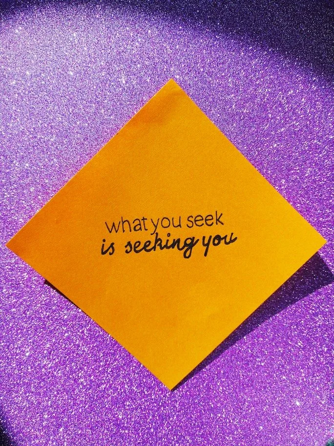Ein Post-it liegt auf einer glitzernden, pinken Fläche. Darauf steht: What you seek, is seeking you.