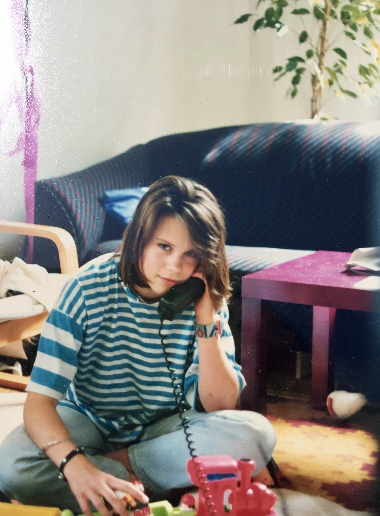 Die Bloggerin Julia von junieundich in den 90ern Jahren sitzt in einem Wohnzimmer und telefoniert. Sie sieht klassisch 90er Jahre aus. Mit Pop-Swatch-Uhr und der typischen Popper-Frisur
