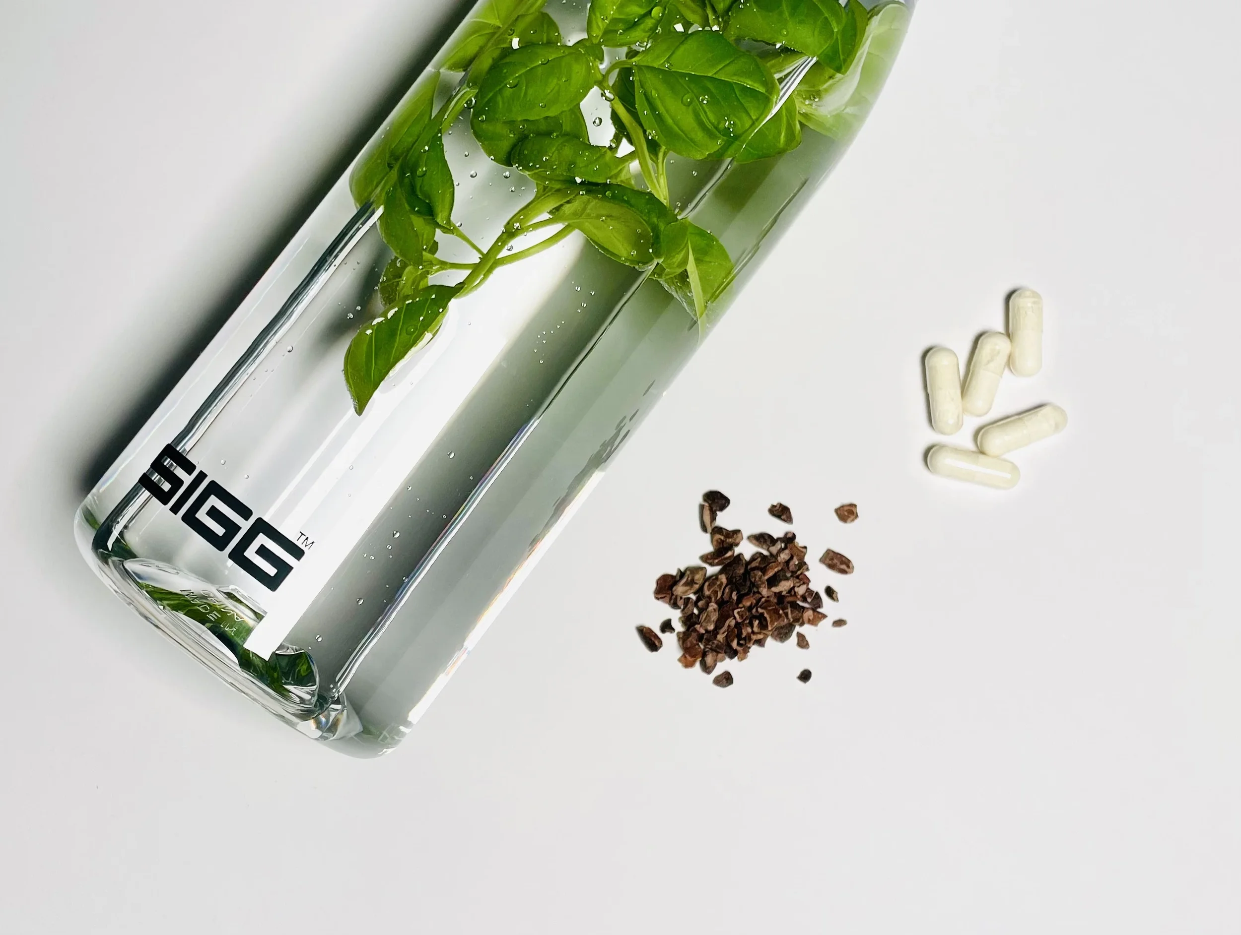 Man sieht eine Trinkflasche von Sigg mit Wasser und Kräutern auf einer weißen Fläche liegen. Daneben liegen Probiotika-Alflorex-Kapseln und Kakaonibs