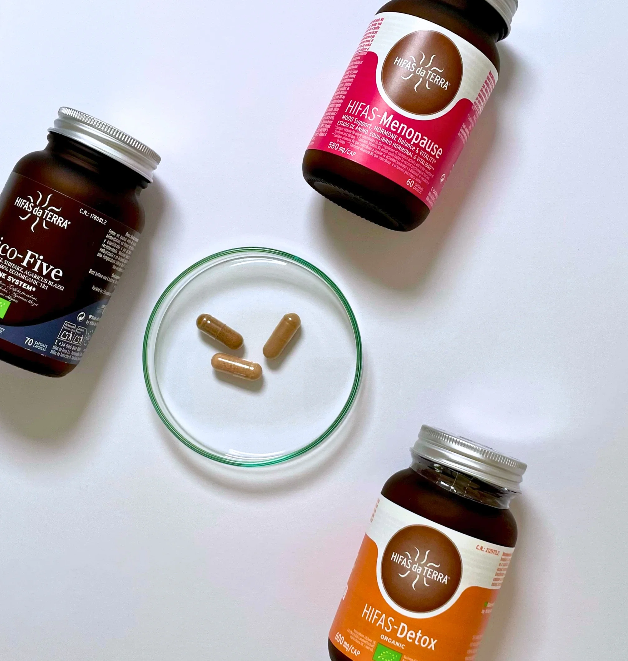 Drei Glasflakons mit Hifas da Terra Produkten, Menopause, Mico-Five und Detox liegen hier, mitten drin liegt eine Petrischale mit drei Kapseln