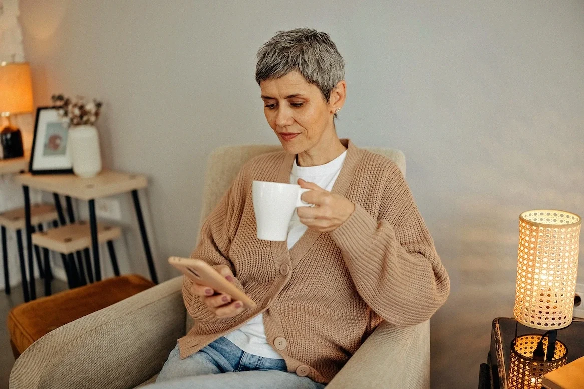 Eine Frau im beigen Cardigan hält eine Tasse in der Hand und schaut auf ihr Handy, wo sie eine Sitzung mit BetterHelp hat