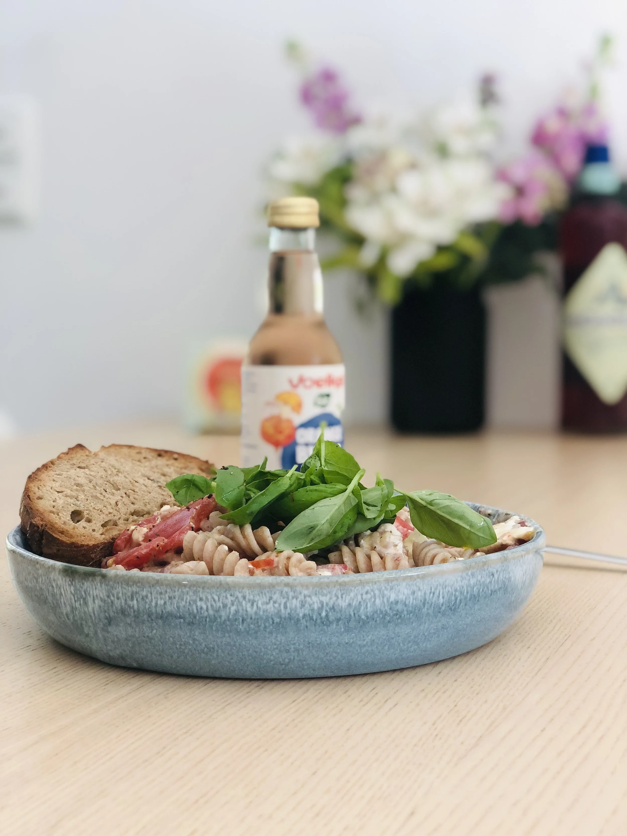 Reisvollkornnudeln in einem blauen Teller, mit frischen Basilikum und einer Scheibe Brot. Im Hintergrund steht eine Flasche London Mule von Voelkel.