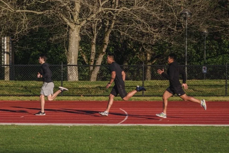 Three young men running on a track field in a park during daytime.
