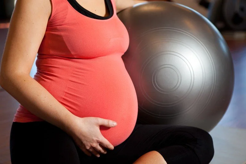 Pregnant woman sitting on the floor with hand on belly in a gym, with an exercise ball in the background.
