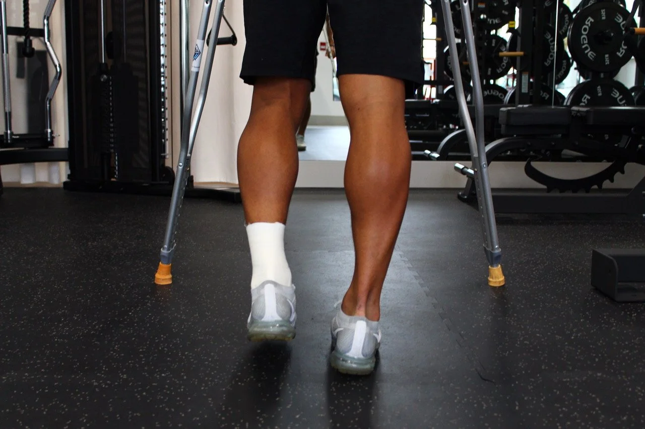 Person walking with crutches, wearing a cast on left foot, in gym setting.