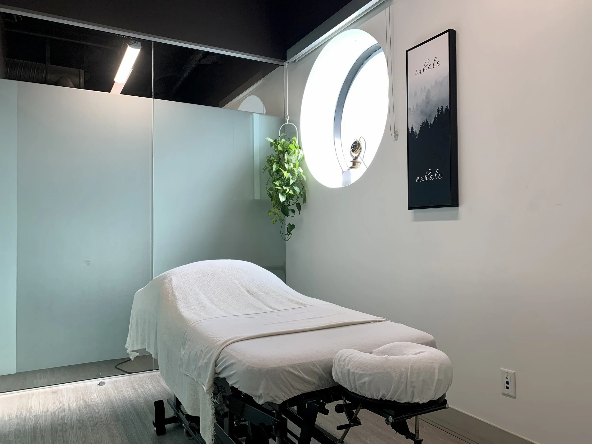 Massage therapy room with massage table covered in white sheet, green hanging plant, oval window with bright light, framed artwork on wall, and white walls.