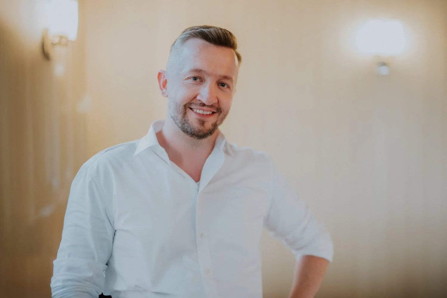 Ein lachender Mann mit blondem Haar und Bart, trägt ein weißes Hemd, steht in einem Raum mit warmem Licht.