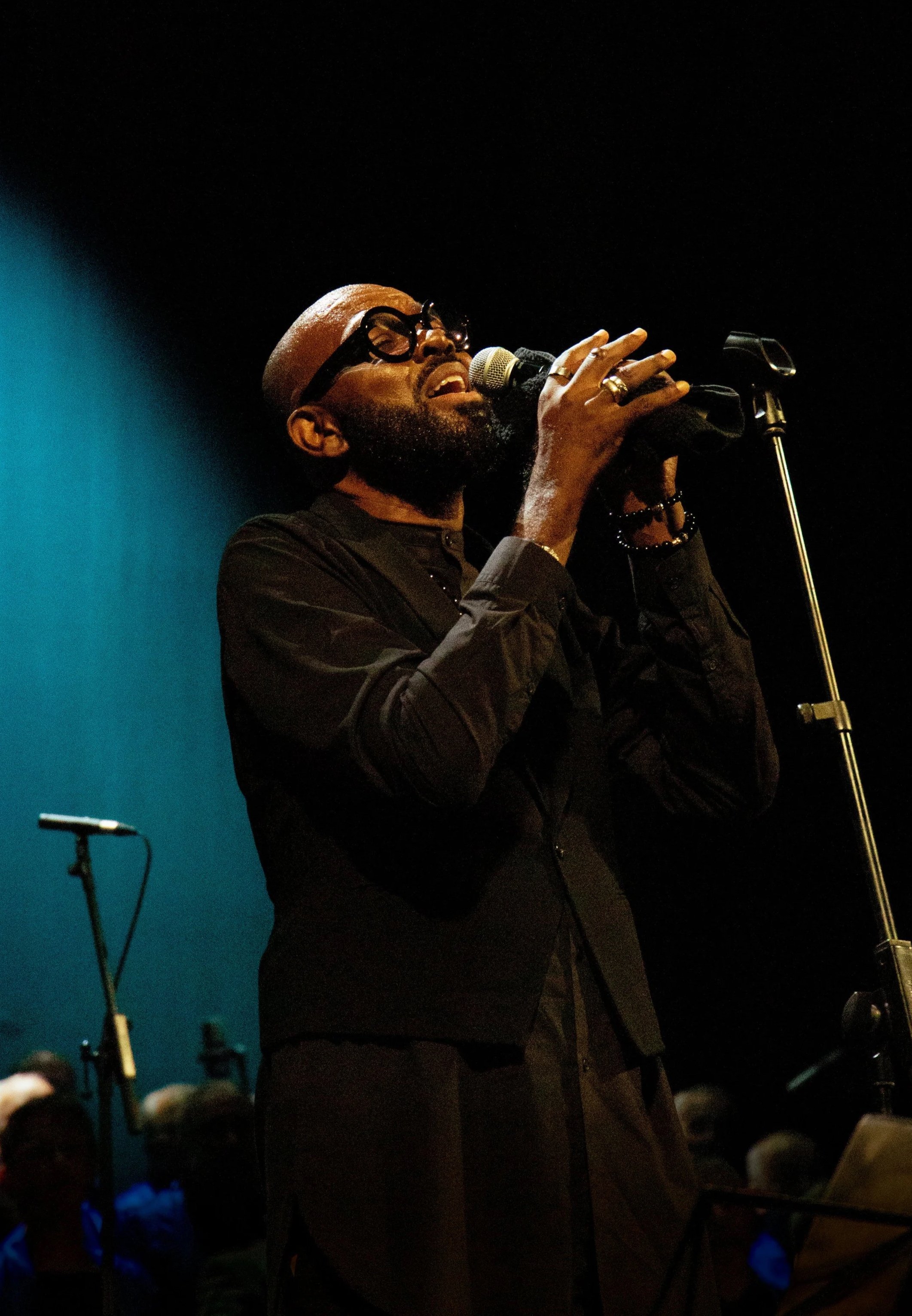 Un hombre negro con barba y gafas, vestido con ropa negra, canta en un micrófono en un escenario oscuro con fondo azul.