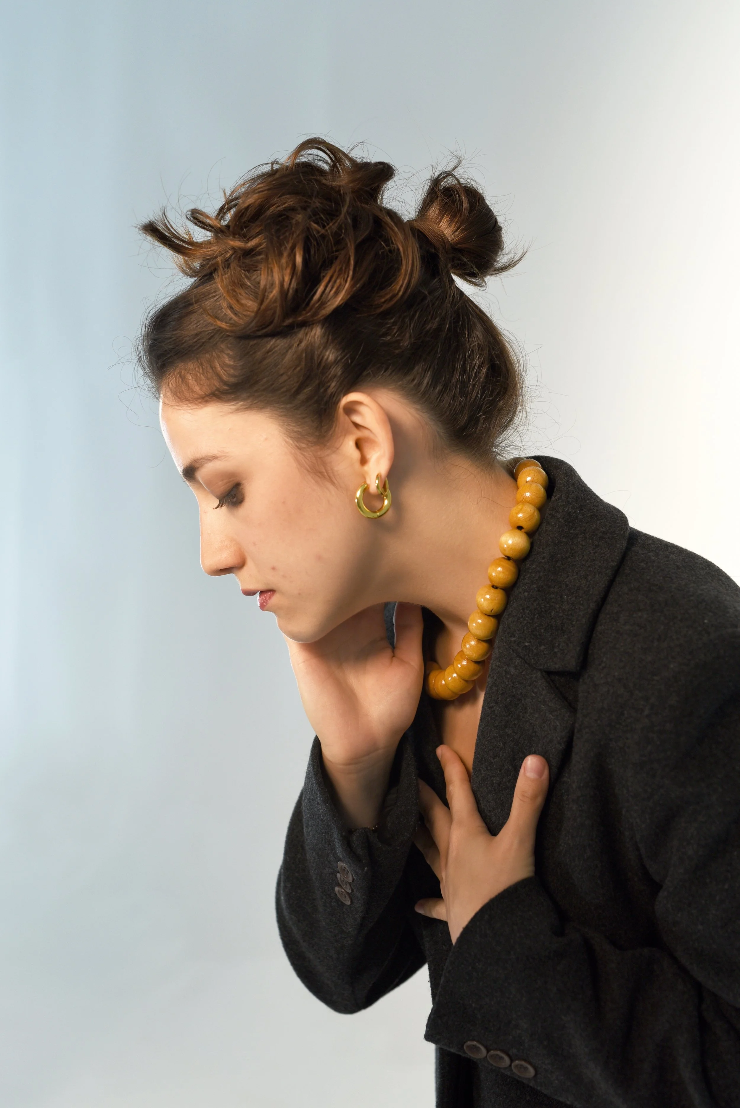 Mujer con cabello peinado en coletas, usando pendientes de aro y collar de madera, vistiendo blazer gris, tocándose el cuello con expresión pensativa.