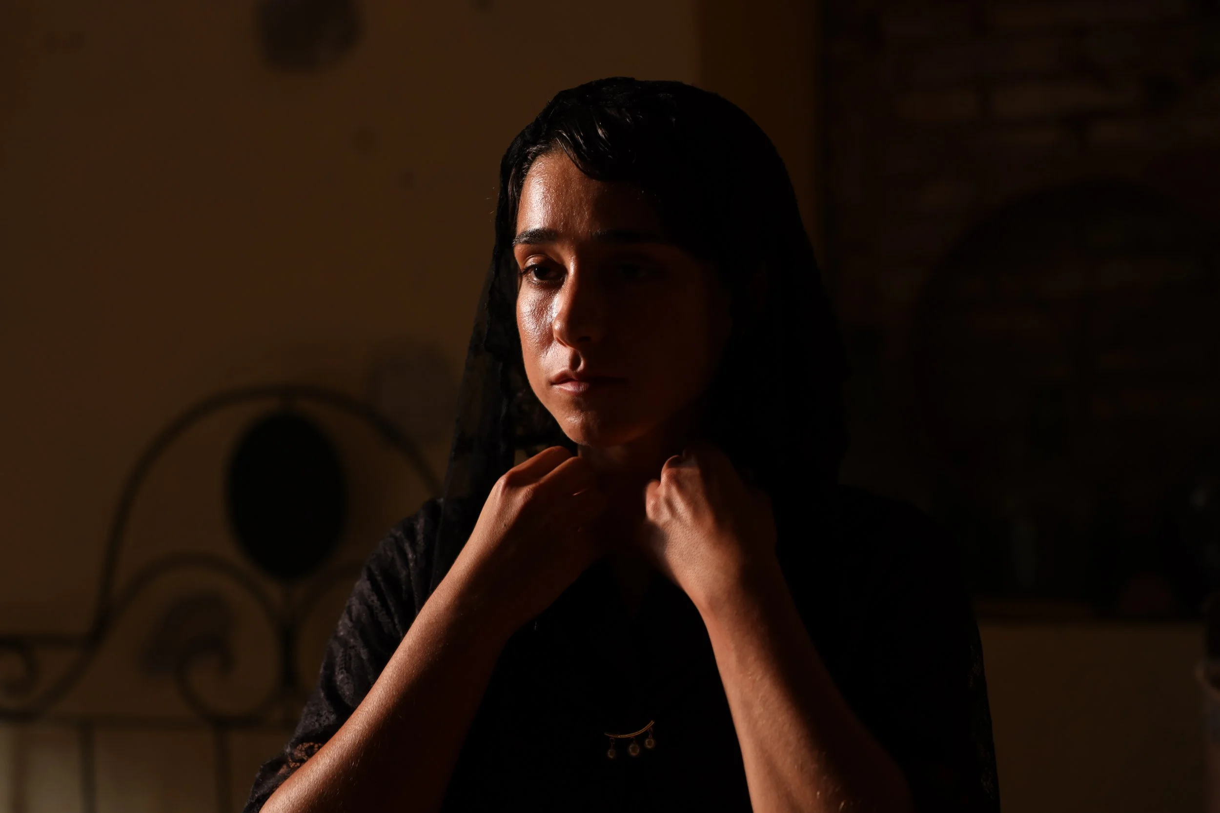 Mujer con vestido negro y velo negro ajustándose el cuello en una habitación con iluminación tenue.