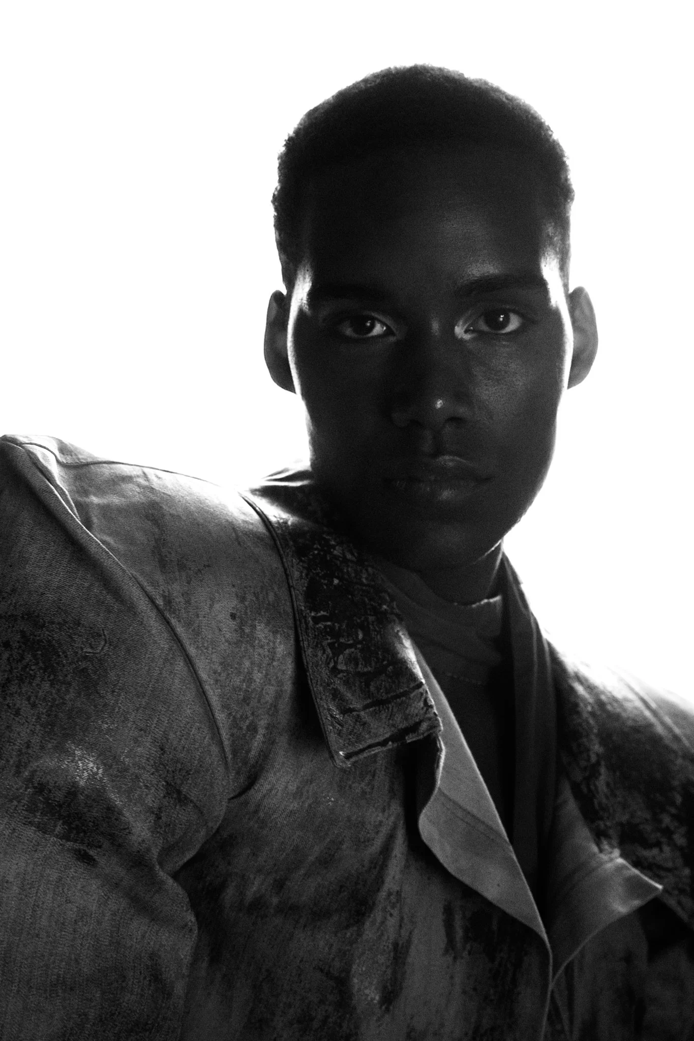 Retrato en blanco y negro de un joven con cabello corto, rostro serio y expresión pensativa, con una chaqueta de mezclilla y fondo blanco brillante.