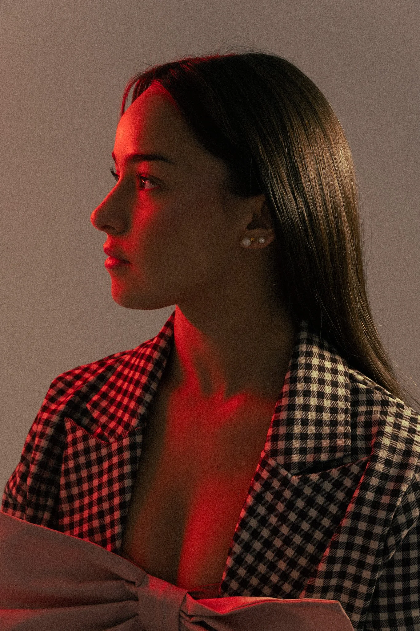 Retrato de una mujer de perfil con cabello largo y oscuro, vestida con una camisa de cuadros y sujetando una prenda blanca, con iluminación roja en la mejilla y el cuello.