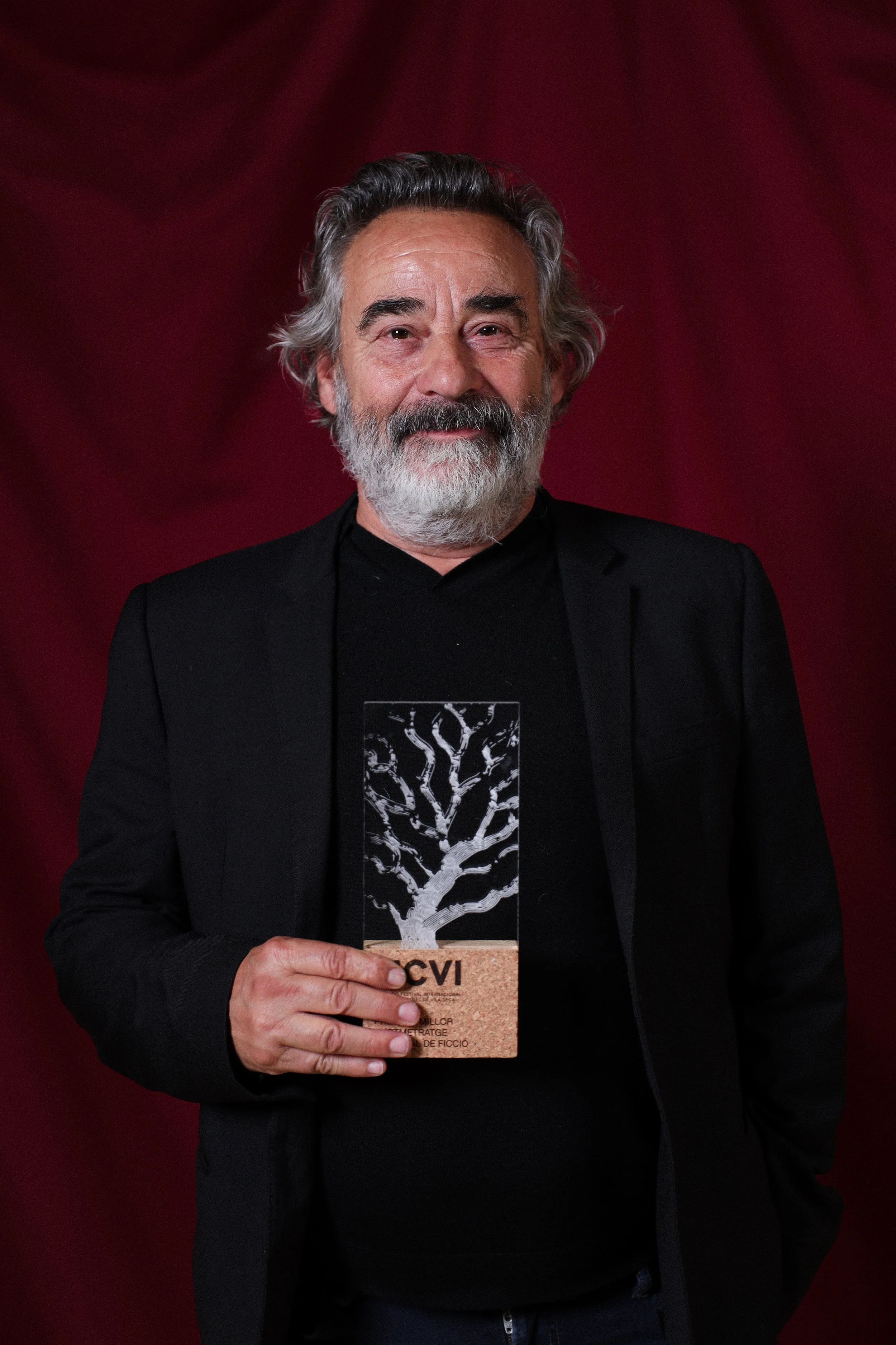 Un hombre de cabello gris, barba y bigote, viste un traje negro y sostiene un premio en un fondo de cortina roja.