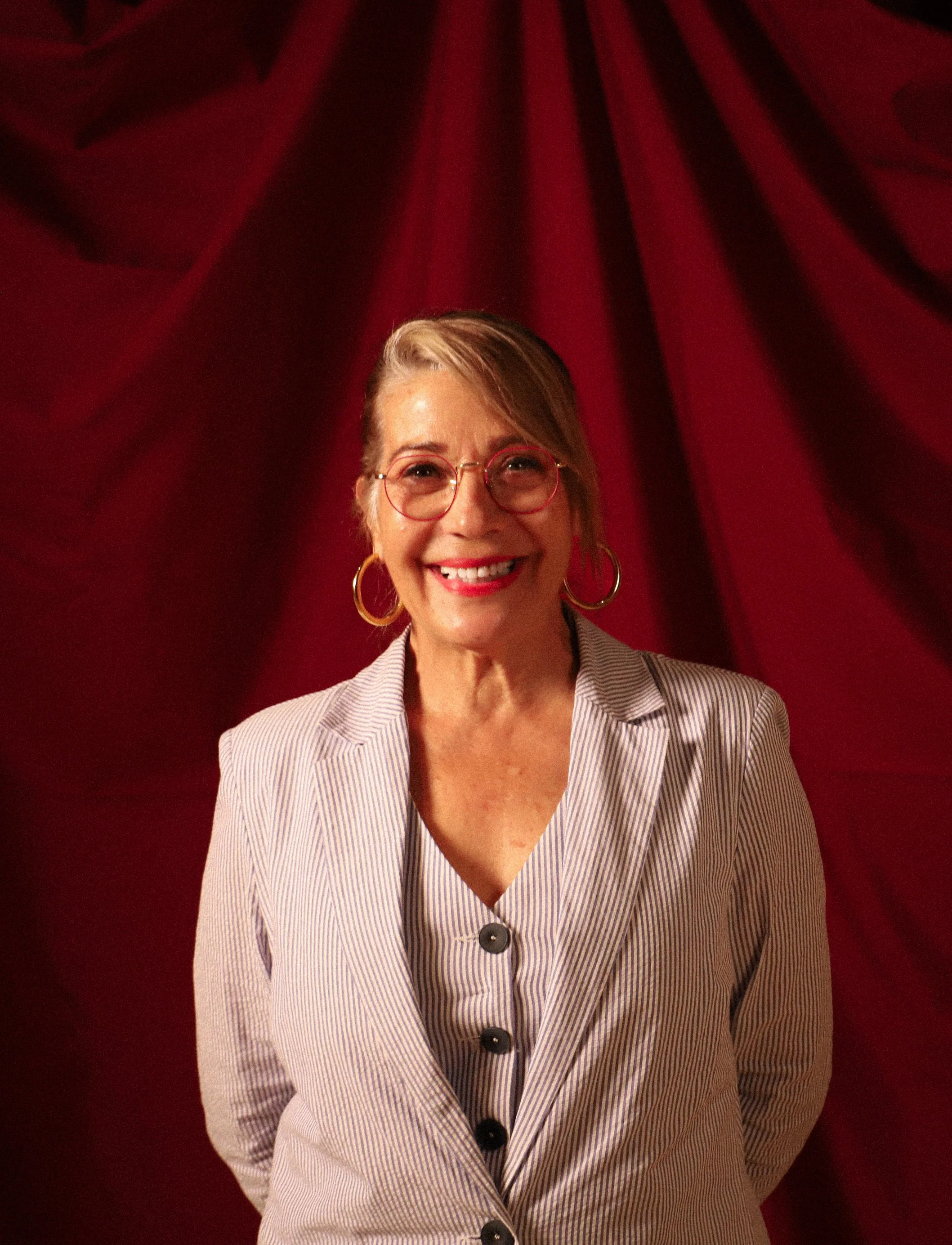 Mujer joven con gafas rojas, bufanda con rayas y grandes aros dorados, sonriendo frente a cortina roja.
