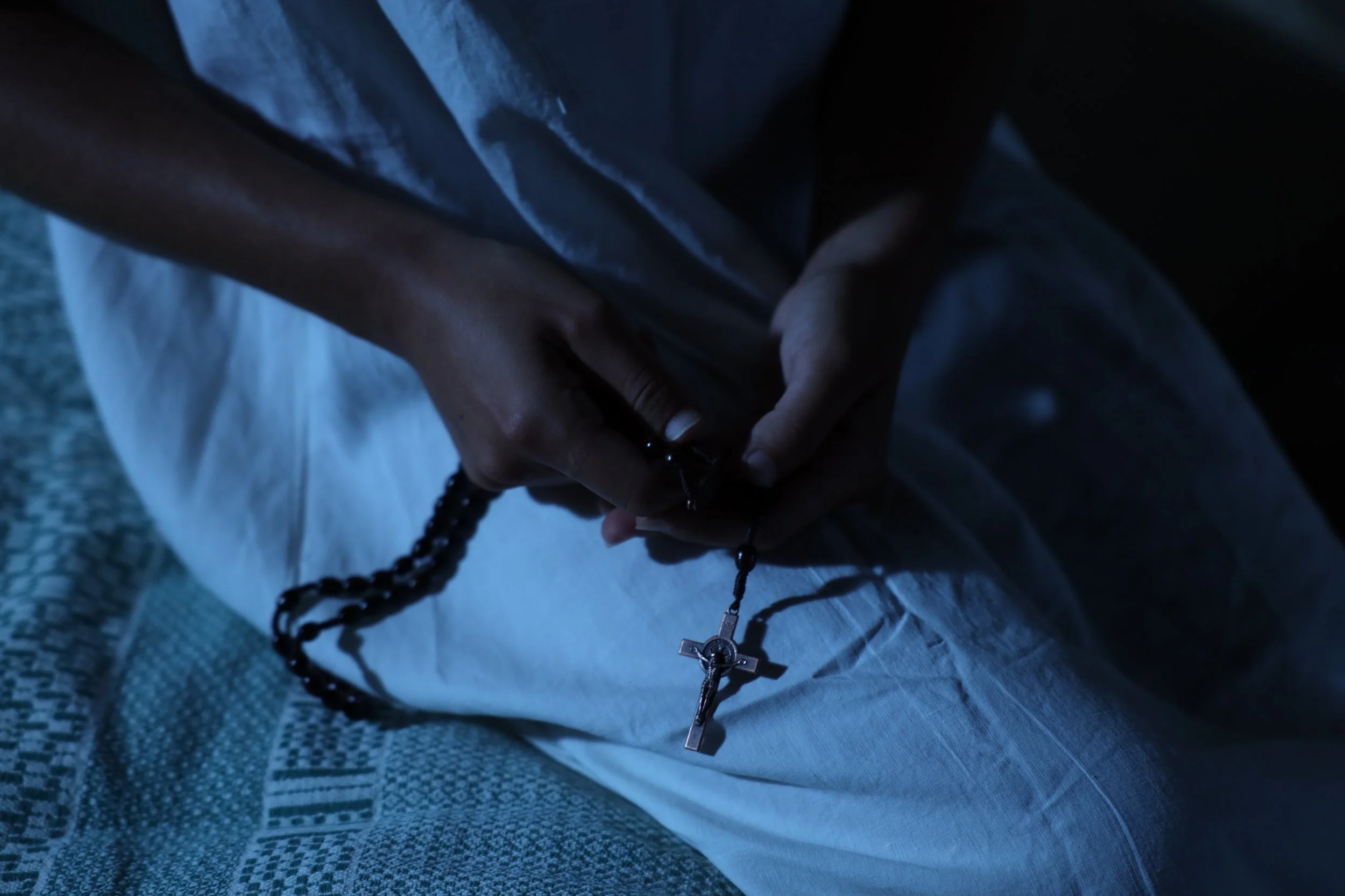 Manos sosteniendo un rosario con una cruz, en un entorno oscuro, con poca iluminación
