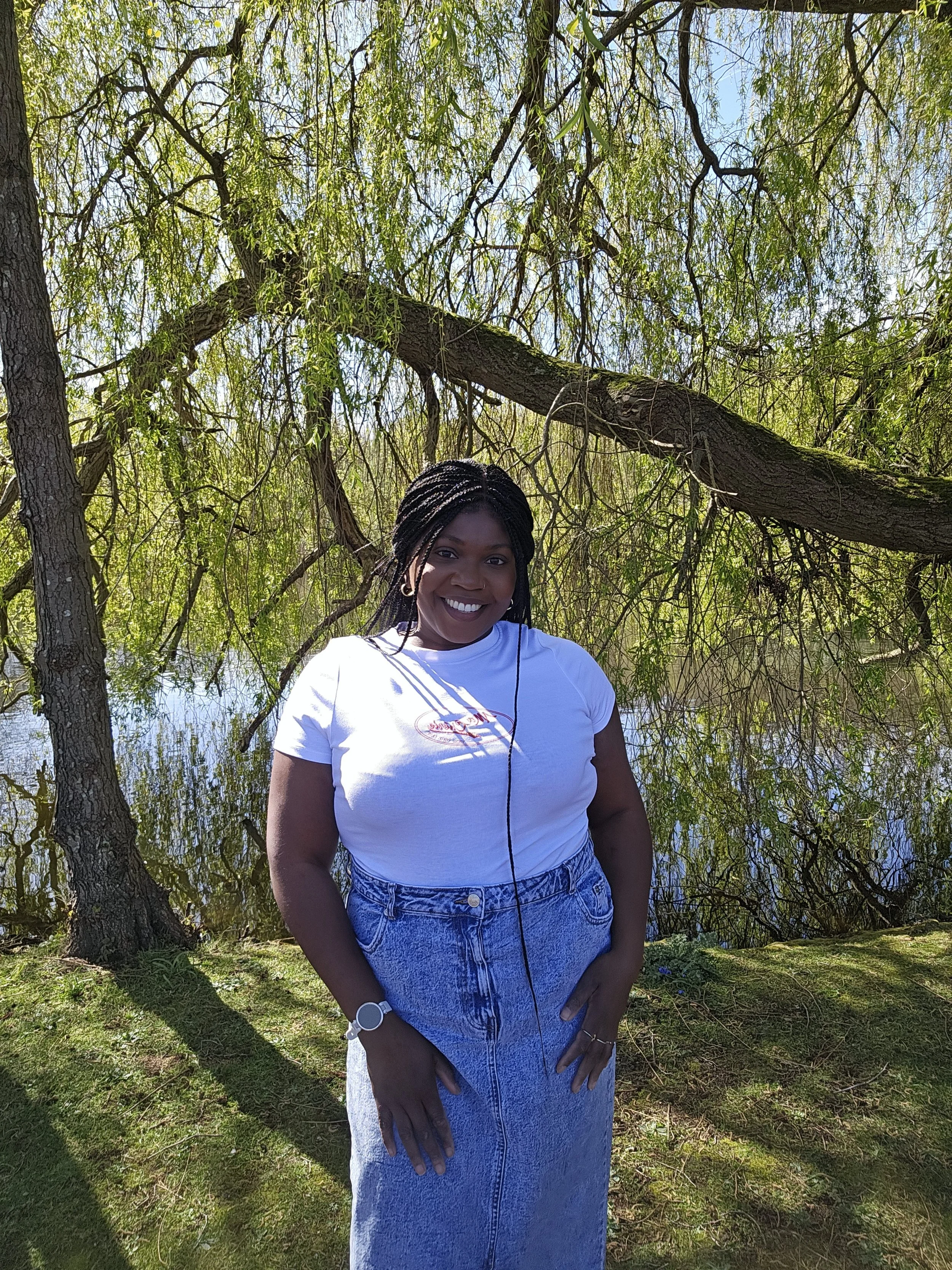 Nicole Vassell, a Black woman with long black braids, smiles in front of a leafy tree
