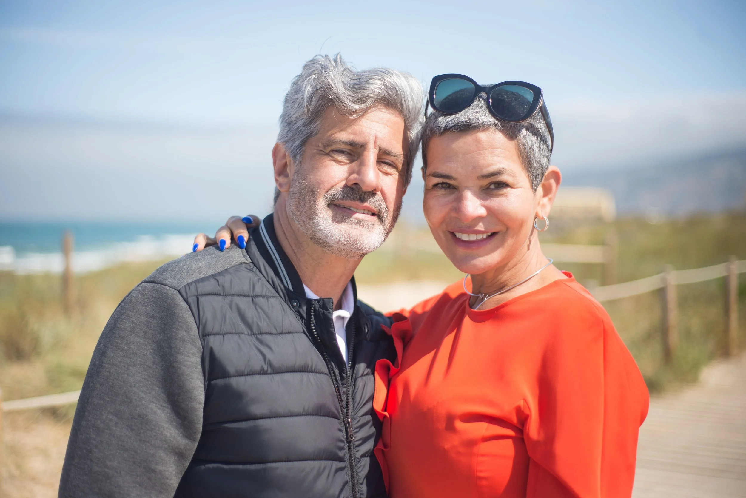 A middle-aged man and woman smiling wth their arms around each other at a beachside location, with the ocean and a cloudy sky in the background.