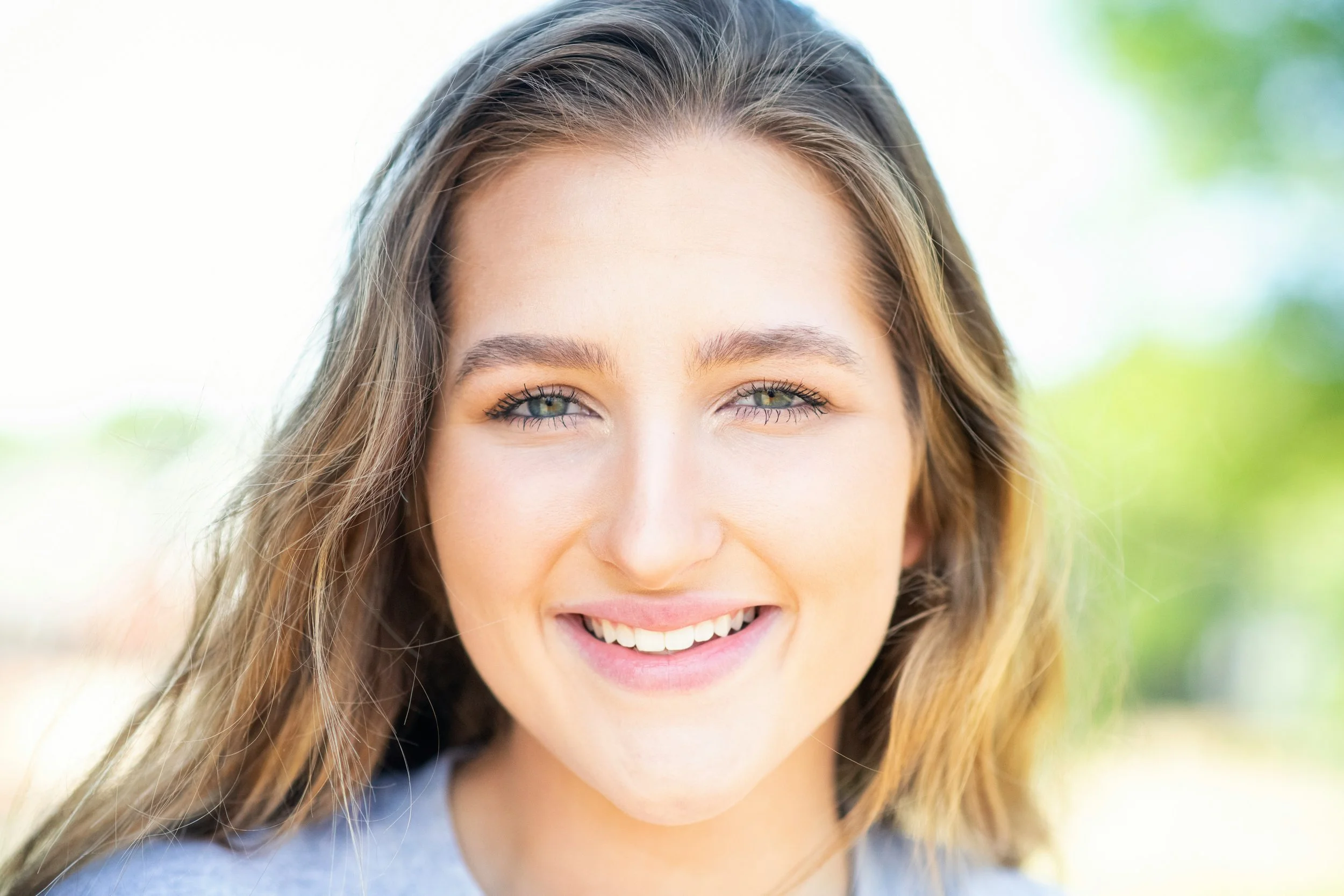 Close-up of a young woman with long, wavy brown hair smiling outdoors, with a blurred green background.