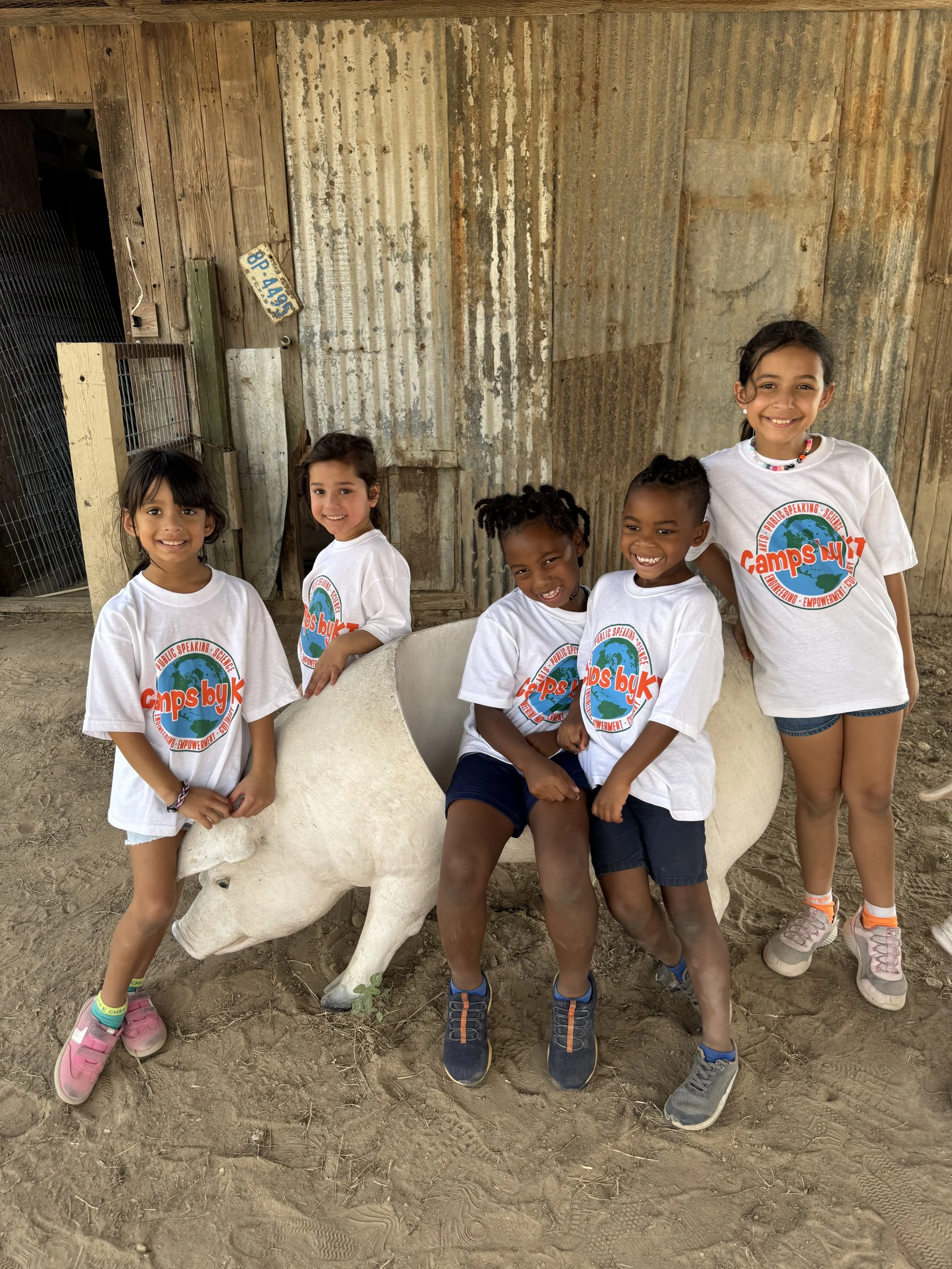 Three smiling young girls standing against an orange wall, wearing colorful casual shirts, with the central girl wearing a pink headband and a cape.