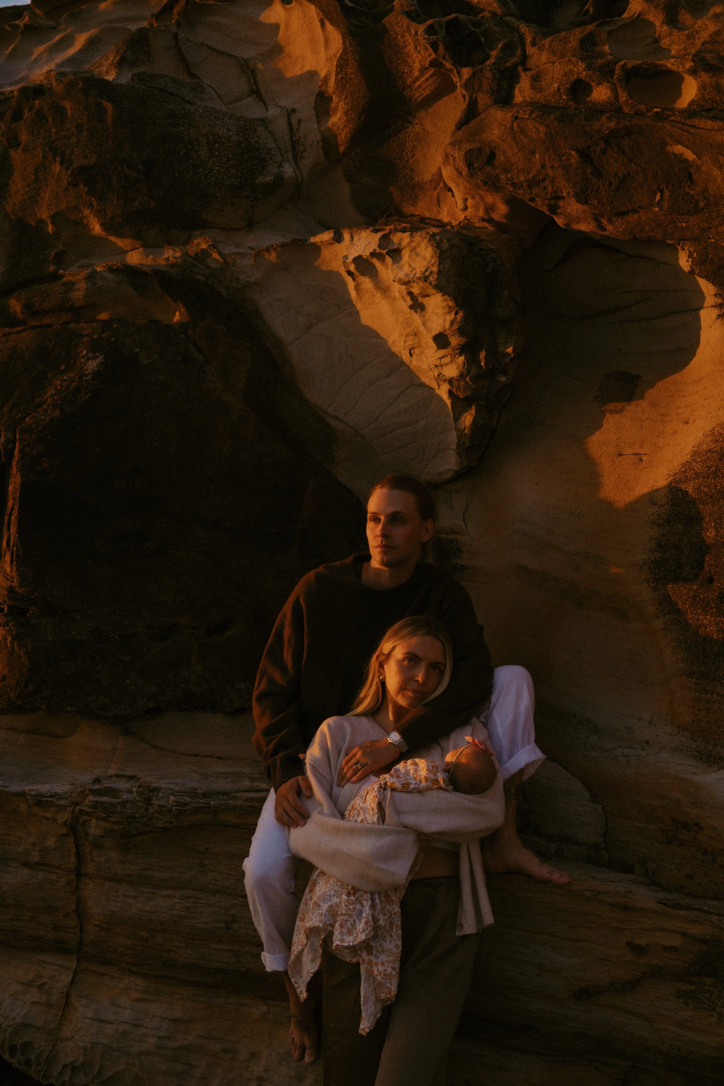 A woman with blonde hair holding a newborn baby wrapped in a blanket, sitting in front of a woman with dark hair and a black sweater, with a rocky background.