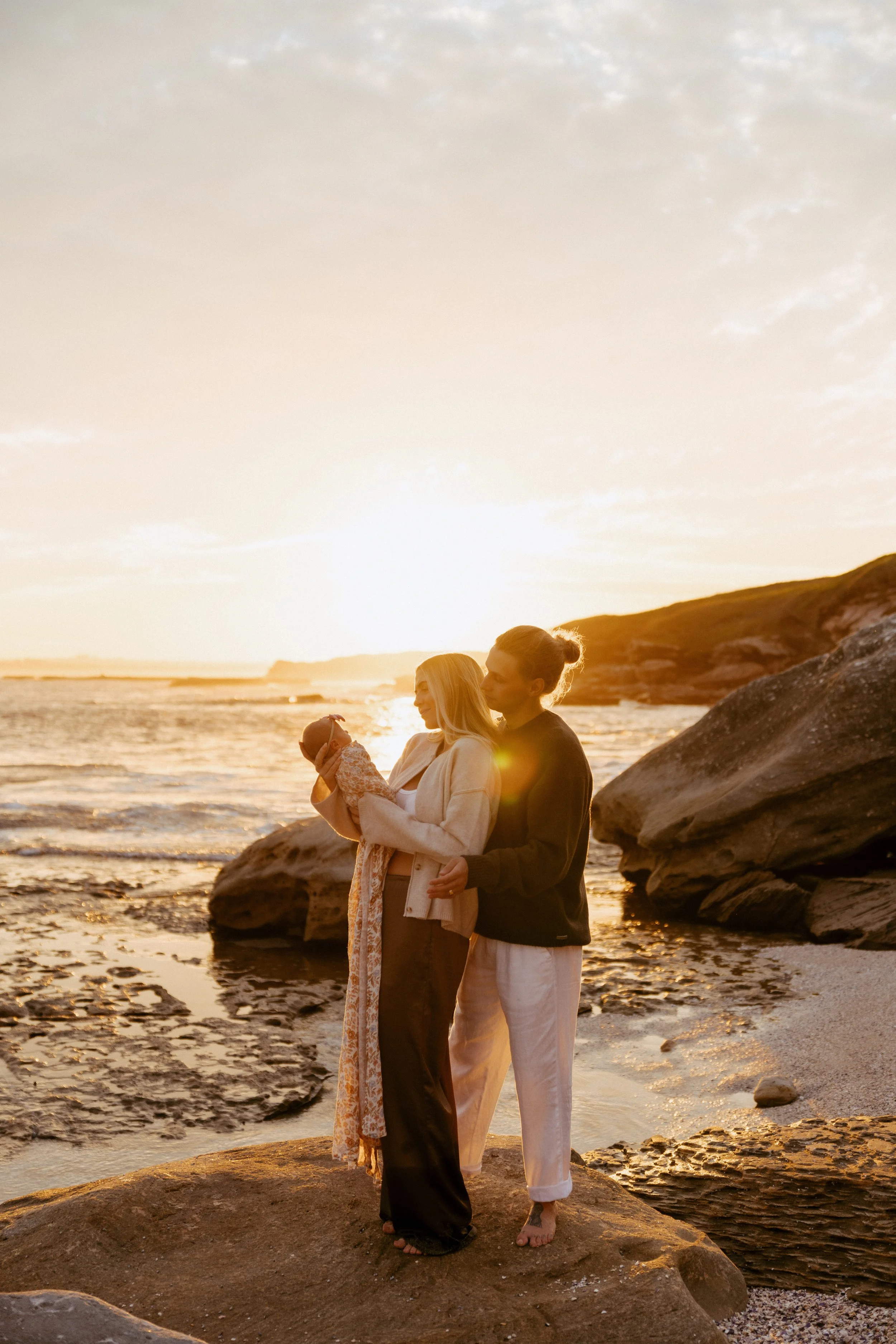 A man and women and a baby on rocks by the ocean during sunset, with cliffs in the background.