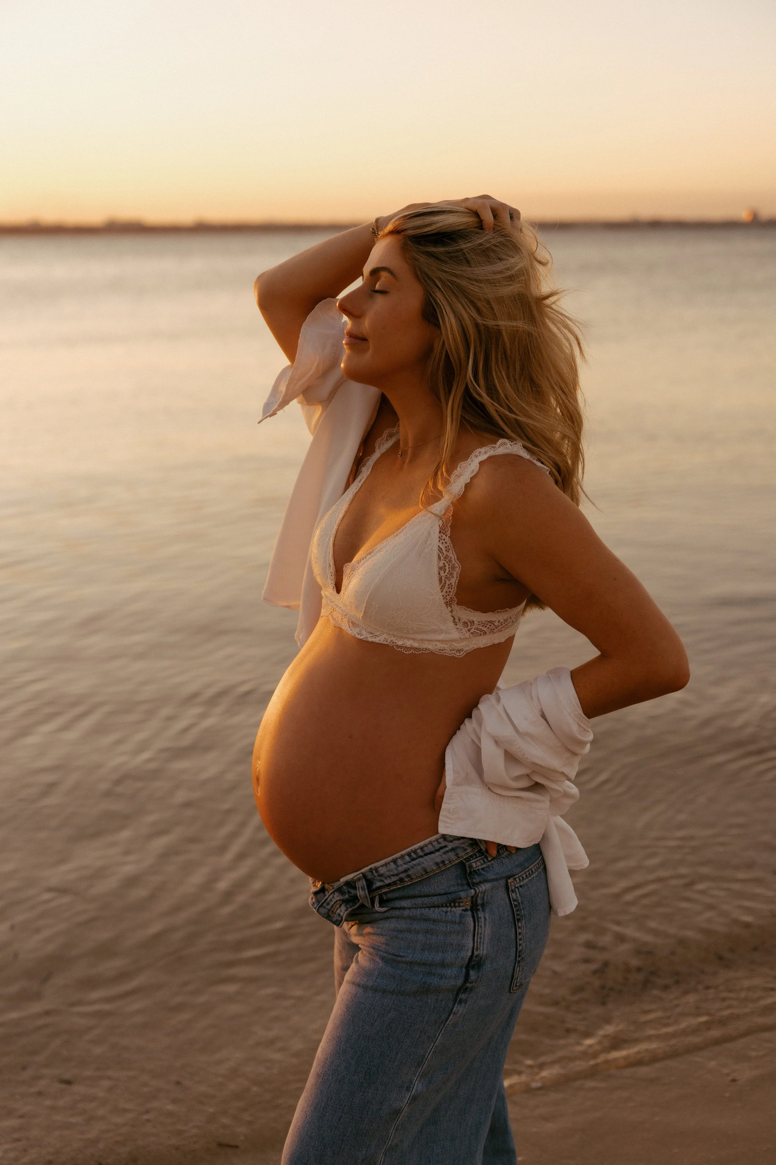 Pregnant woman standing by the water at sunset, wearing a white lace crop top and jeans, with her eyes closed and a peaceful expression.