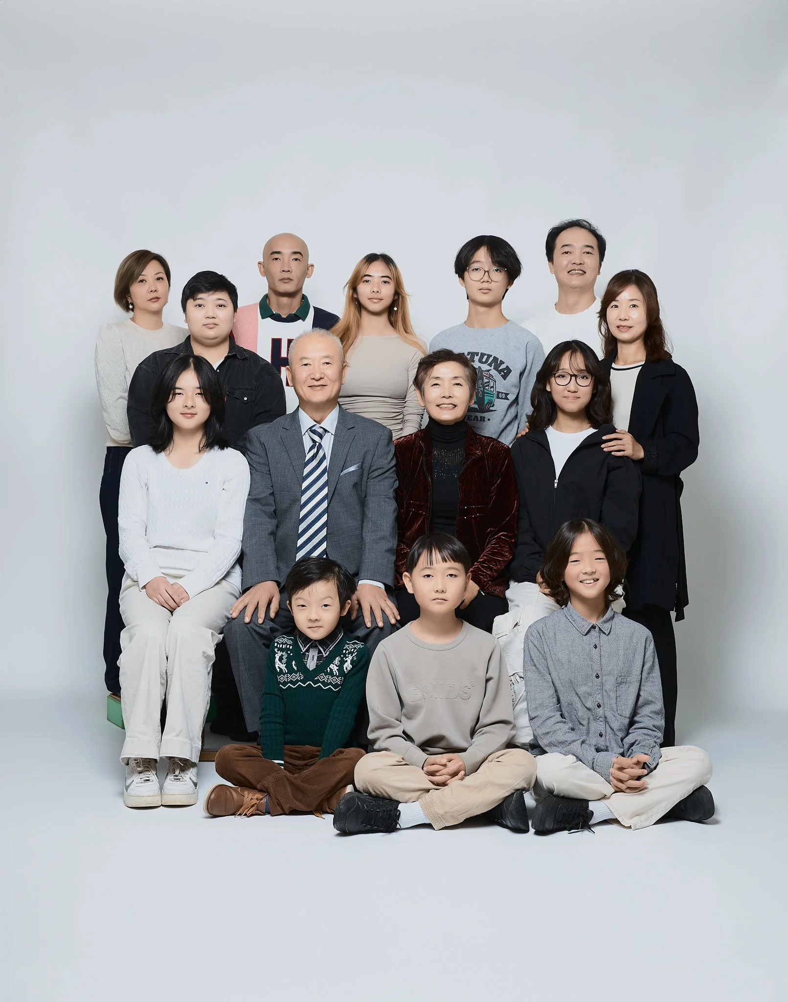 A multigenerational family portrait with adults and children sitting and standing against a plain background. The family includes both men and women, ranging from young children to elderly adults. They are dressed in casual to semi-formal outfits.