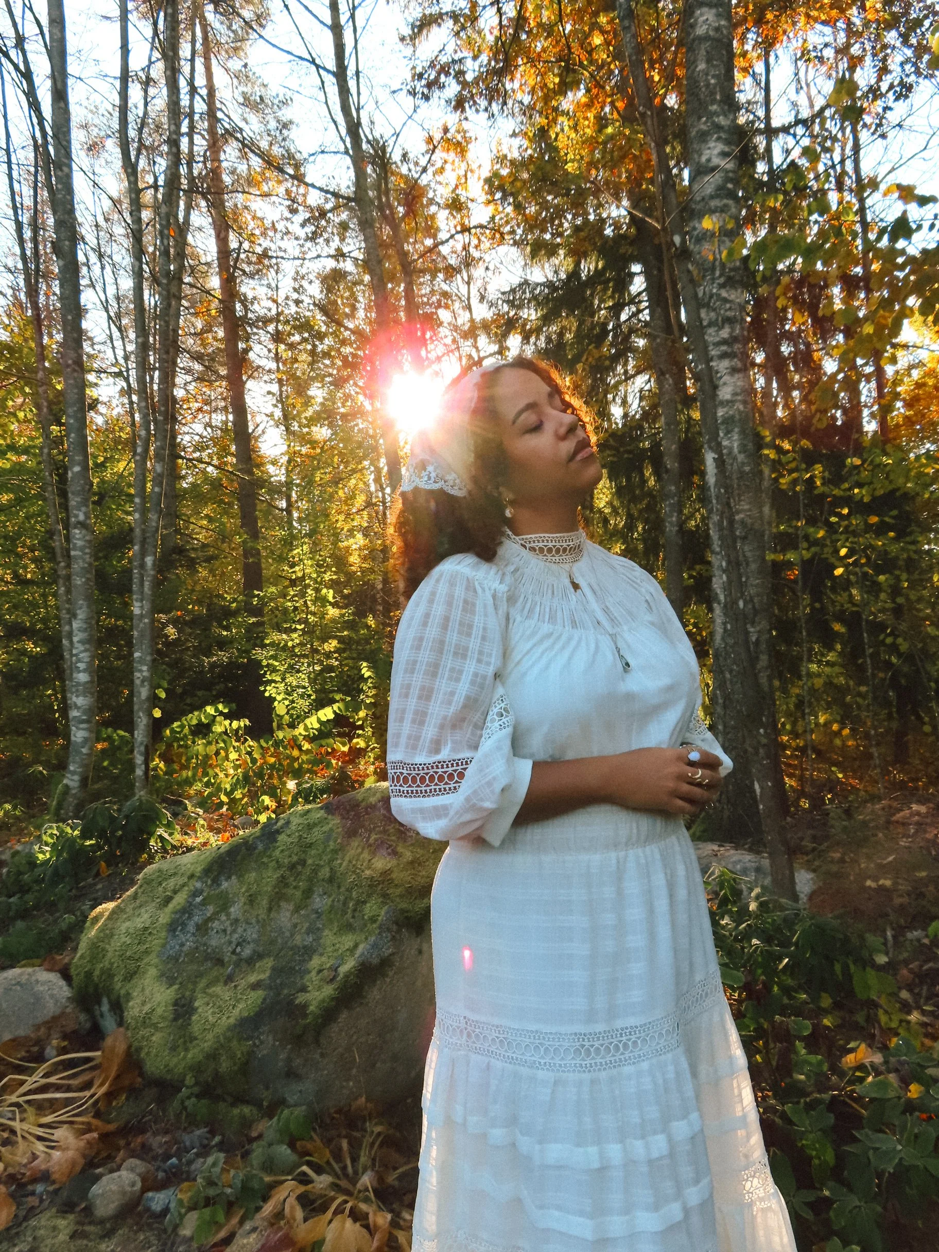 A woman in a white vintage-style dress with lace details, standing in a forest at sunset with her eyes closed, sunlight shining behind her, surrounded by trees and moss-covered rocks.