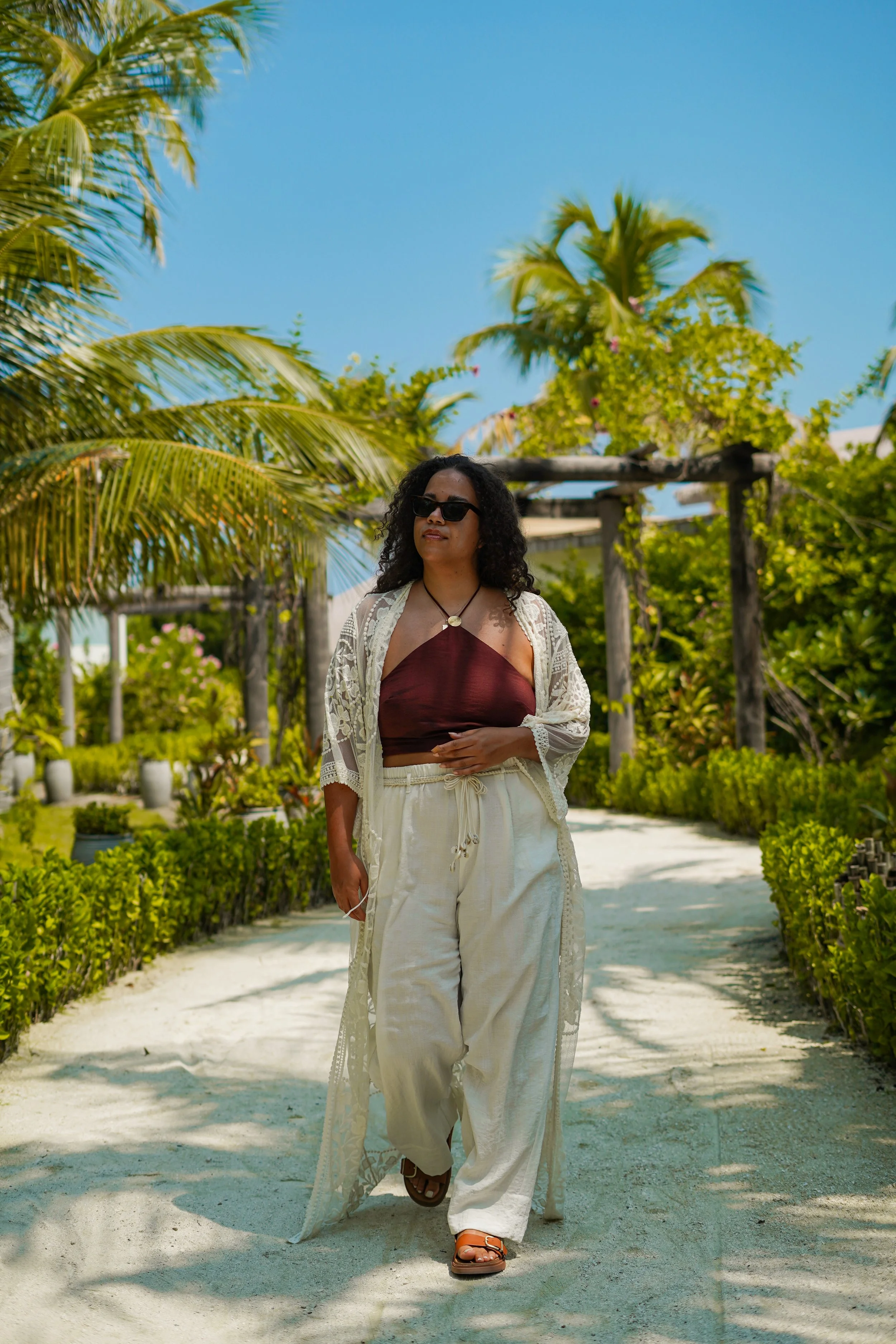 A woman wearing sunglasses, a brown crop top, and white pants, walking along a sandy path in a tropical garden with lush greenery and palm trees under a clear blue sky.