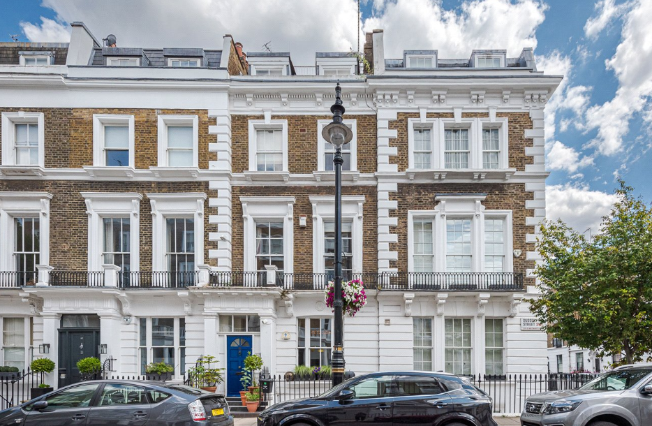 Street view of ornate Victorian-style terraced buildings with white decorative stonework, tall sash windows, and a central lamppost with hanging flowers, with parked cars lining the road.