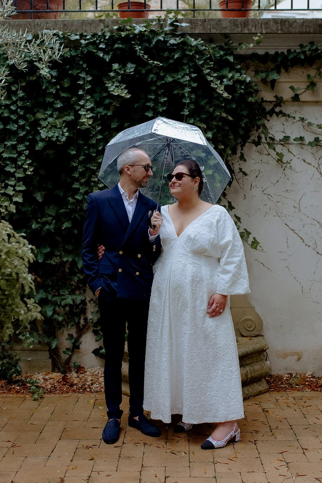 couple de mariés debout sous un parapluie devant un mur de lierres