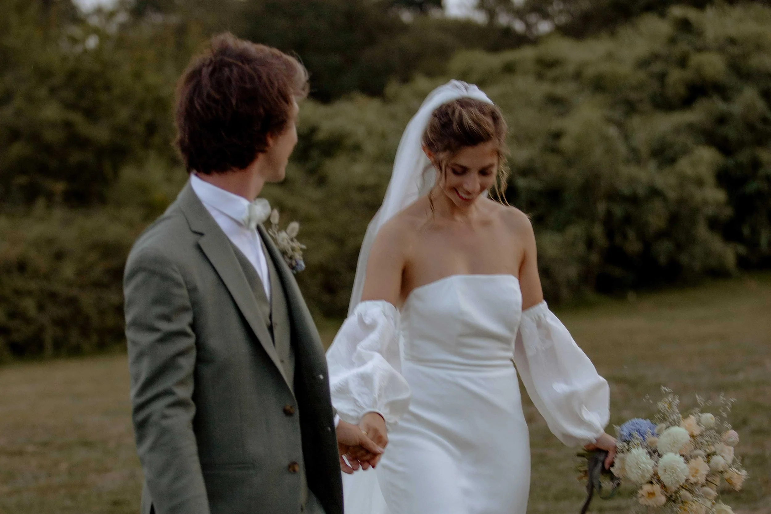 couple de jeunes mariés qui marchent main dans la main dans un champ en riant, le marié regarde vers la mariée qui sourit