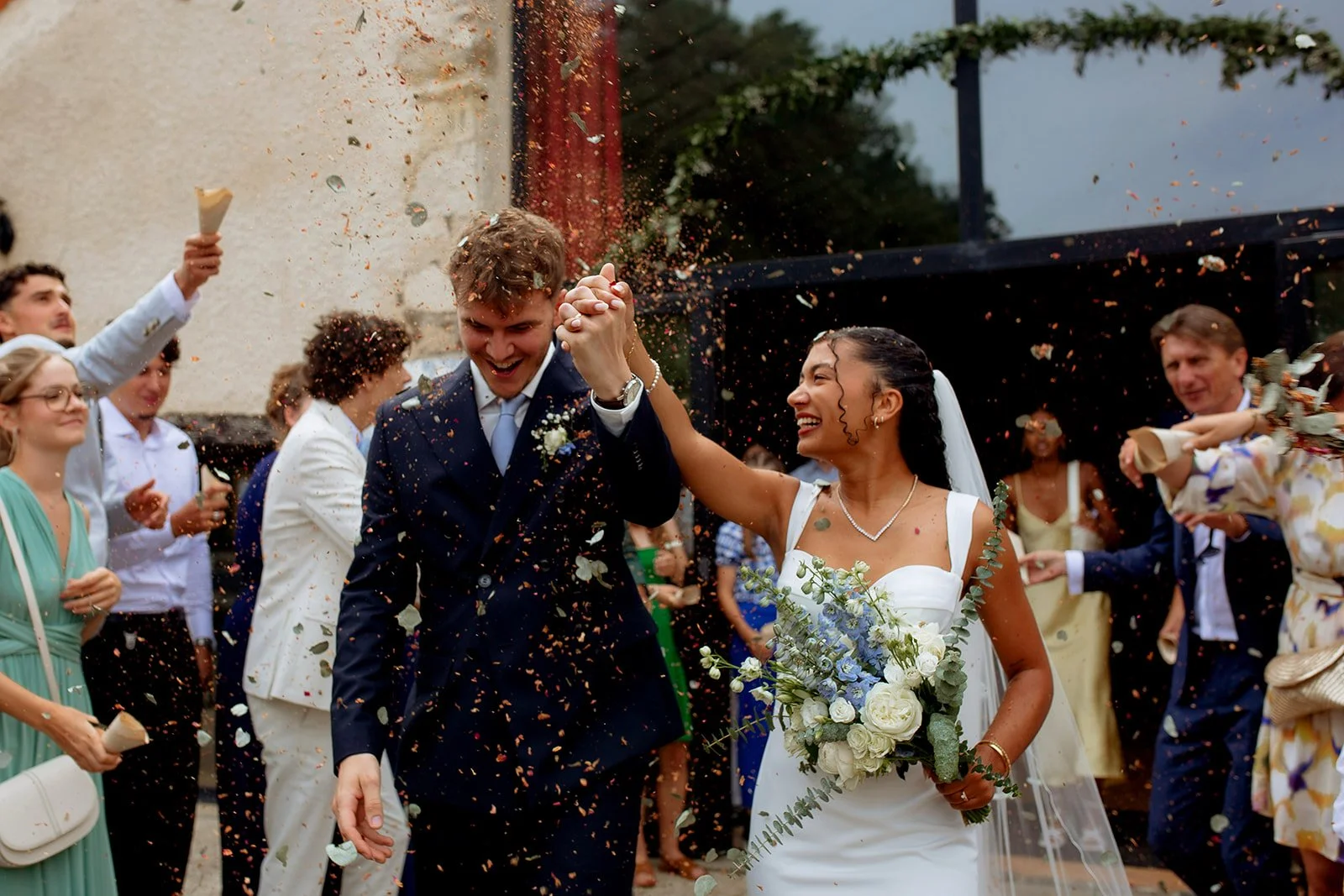 couple de mariés qui sortent de la cérémonie et qui lèvent leurs mains pendant qu'on leur jette des fleurs