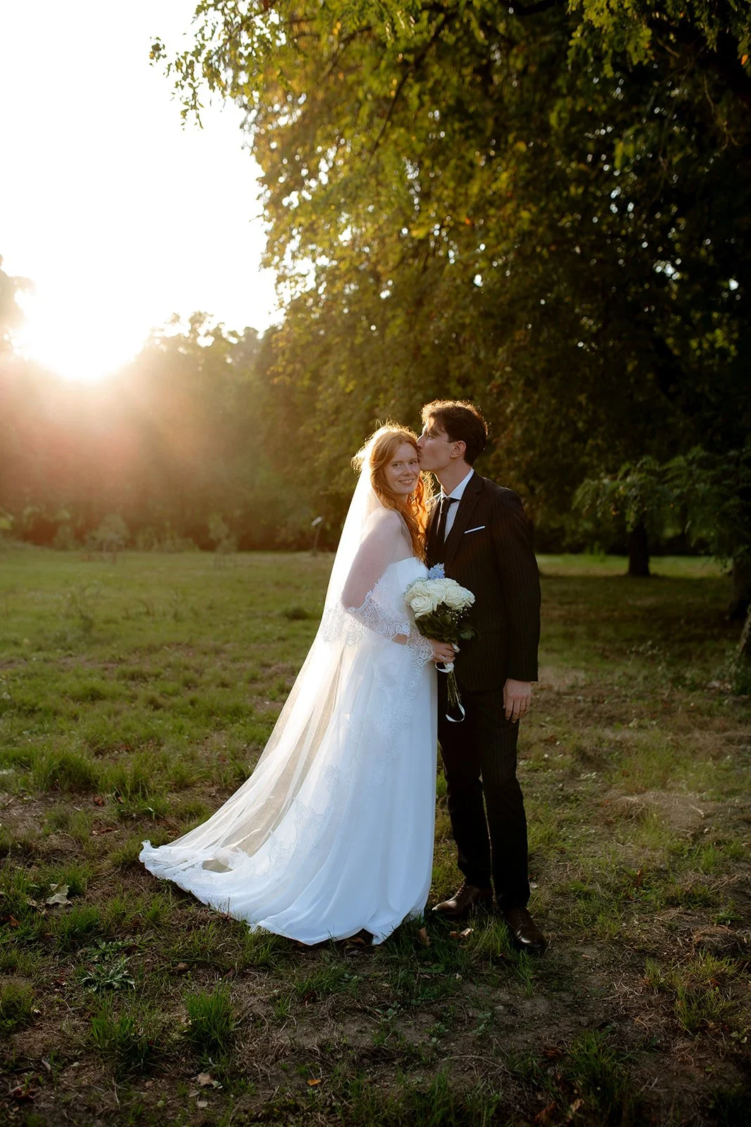 couple de mariés debout dans un champ au coucher du soleil, le marié embrasse la tempe de la mariée qui regarde l'objectif en souriant