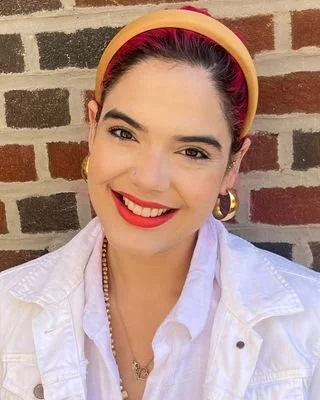 Close-up portrait of a smiling person with dark hair pulled back under a wide yellow headband, wearing red lipstick, gold hoop earrings, and a white collared shirt, standing in front of a brick wall.