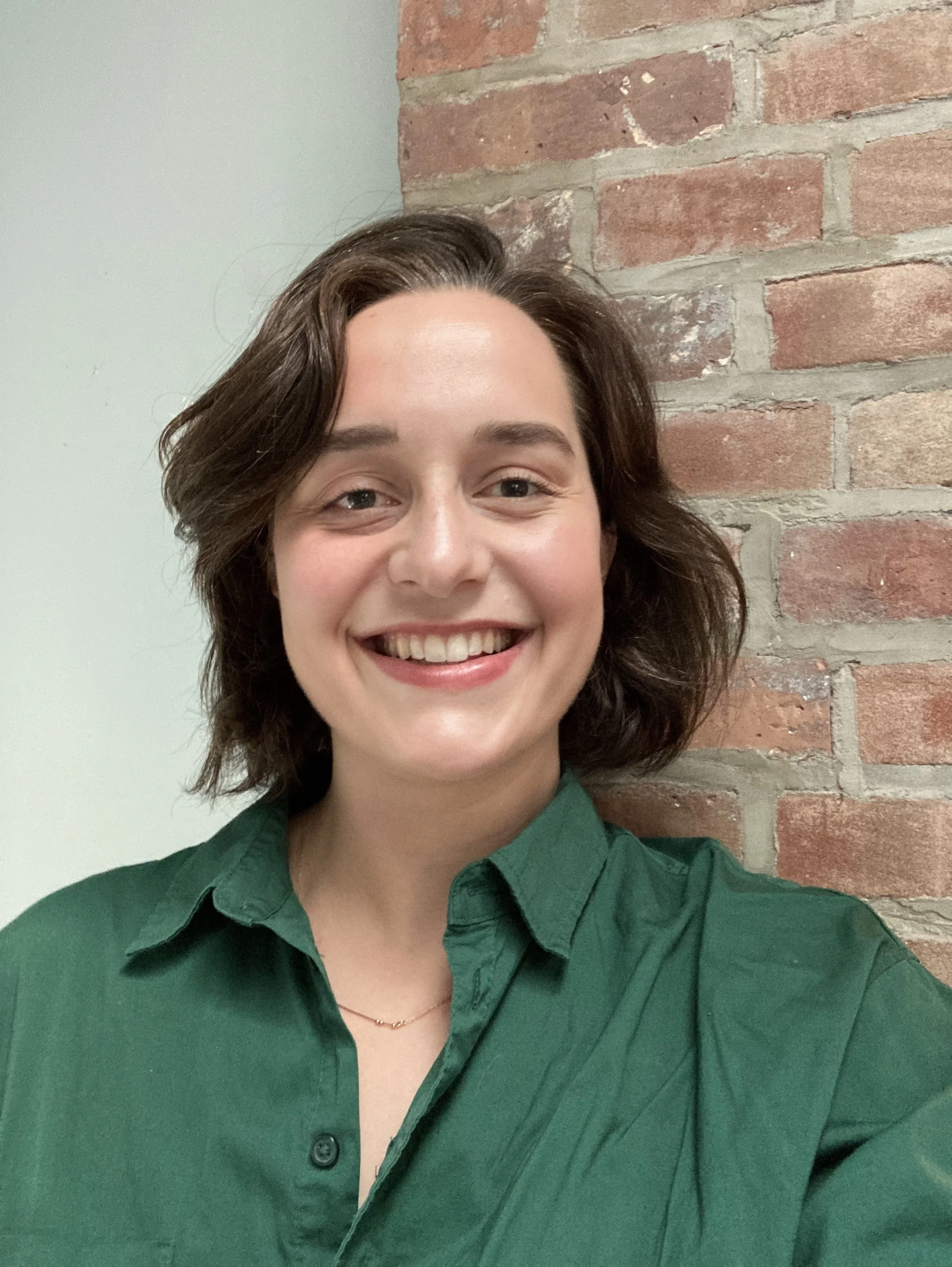 A smiling woman with short dark brown hair wearing a forest green button-up shirt and a delicate necklace standing in front of an exposed brick wall and a light gray wall.