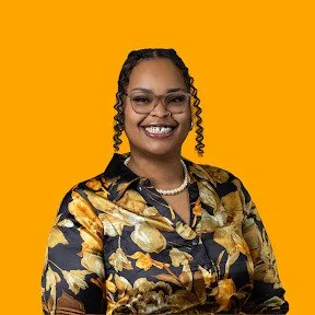 Smiling woman wearing glasses, a pearl necklace, and a black and gold floral blouse, with styled curls framing her face, set against a bright orange background.