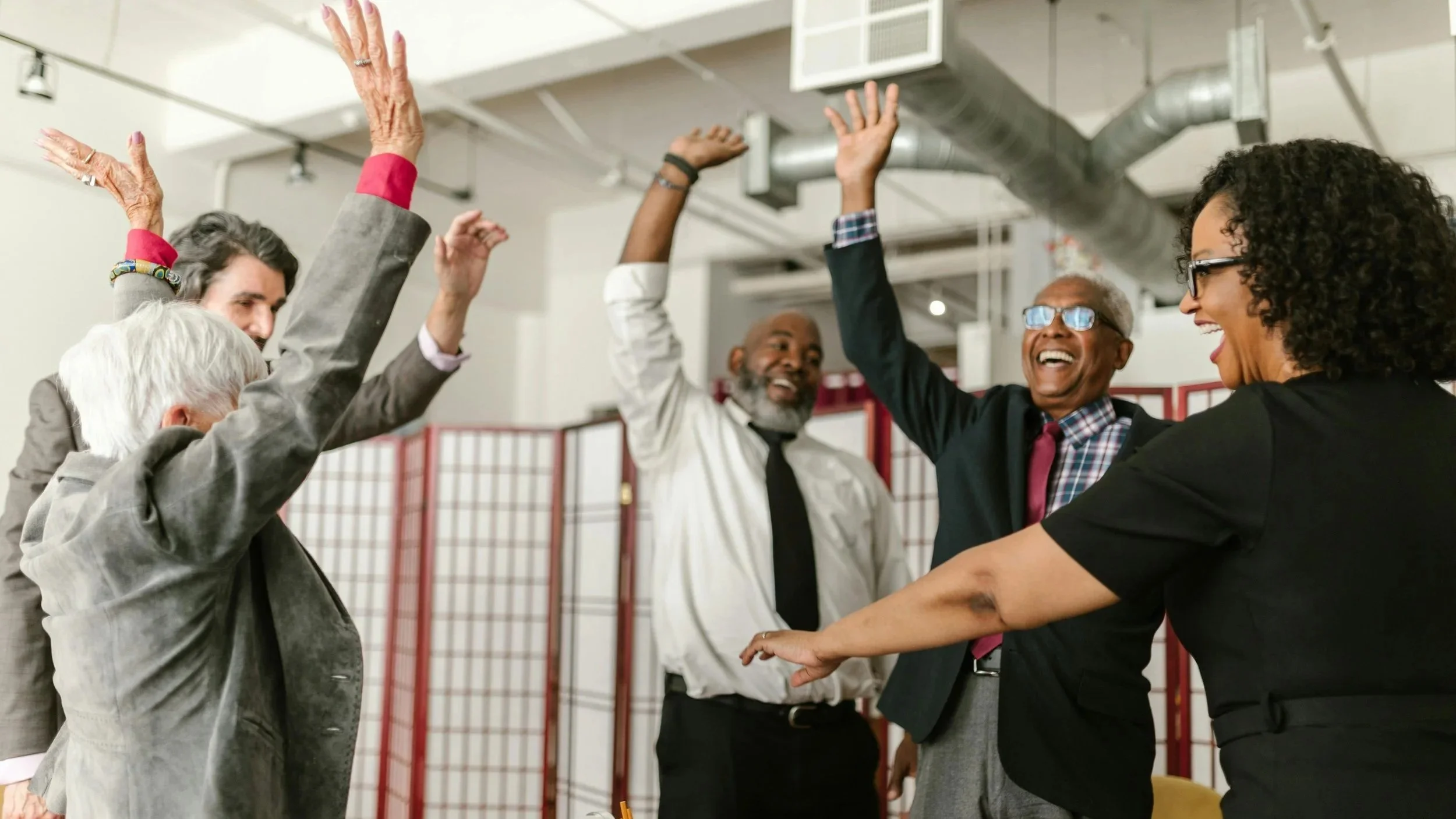 Group of diverse professionals raising their hands together in a celebratory gesture around a table in a modern office space.