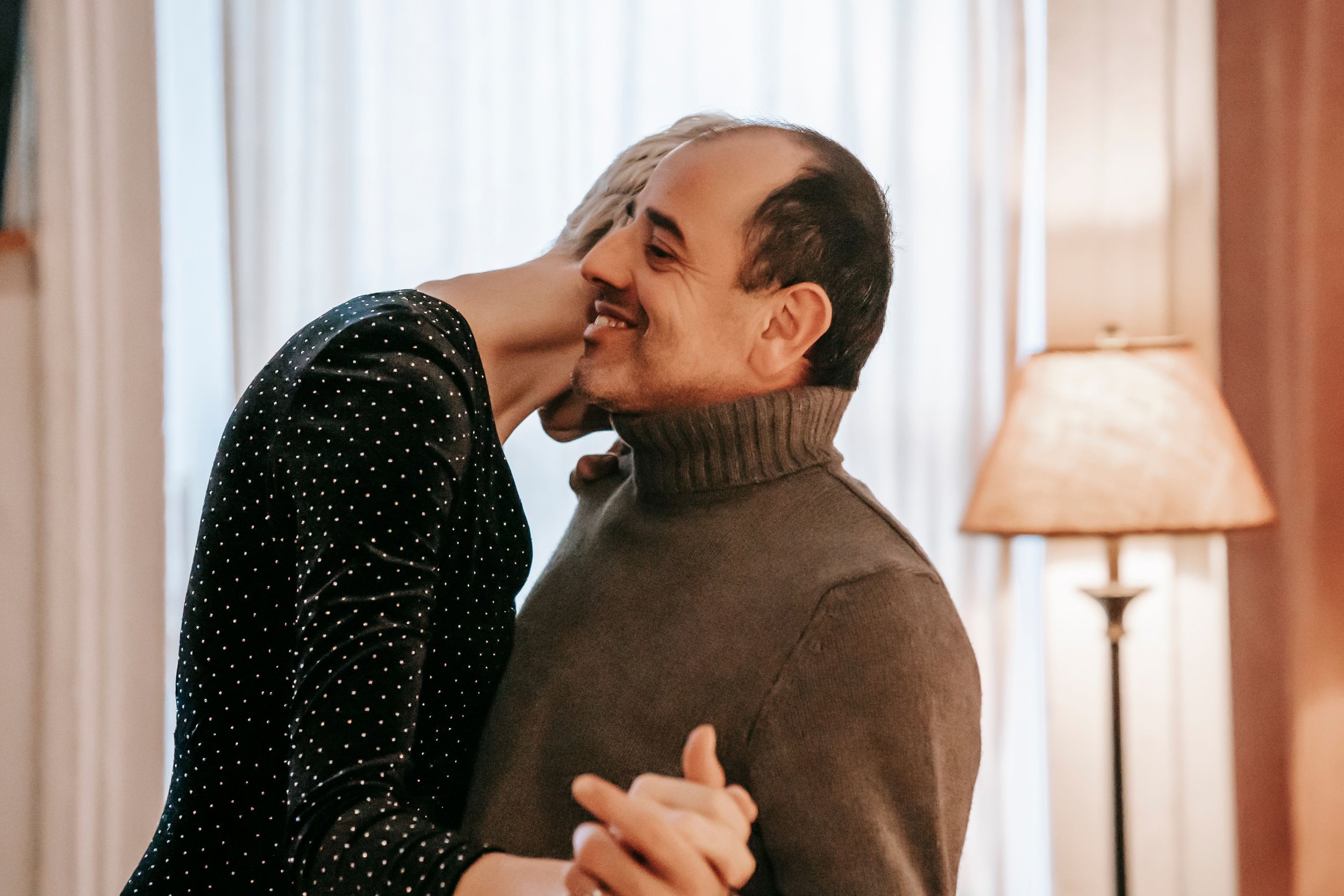 A middle-aged couple slow dances in a softly lit living room, hands clasped, as she presses her forehead on his shoulder while he smiles.