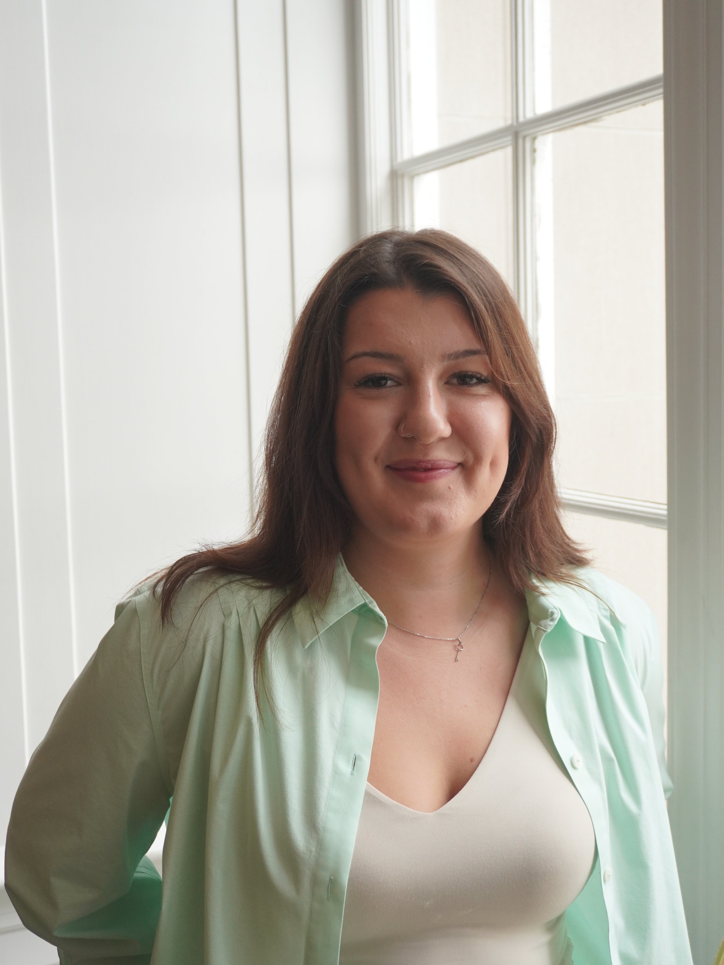Headshot of Defne Sagdic, with a soft, closed-mouth smile and long straight brown hair, wearing a delicate pendant necklace and a mint green button-up shirt over a light top, in front of a white wall and a white-framed window.
