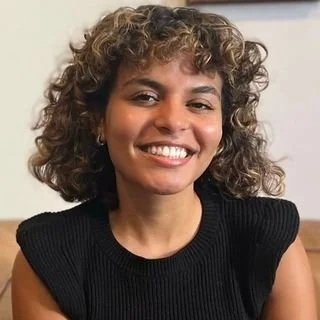 A smiling person with short, curly hair sits indoors, wearing a sleeveless black knit top, with a warm, relaxed expression and a softly lit background.