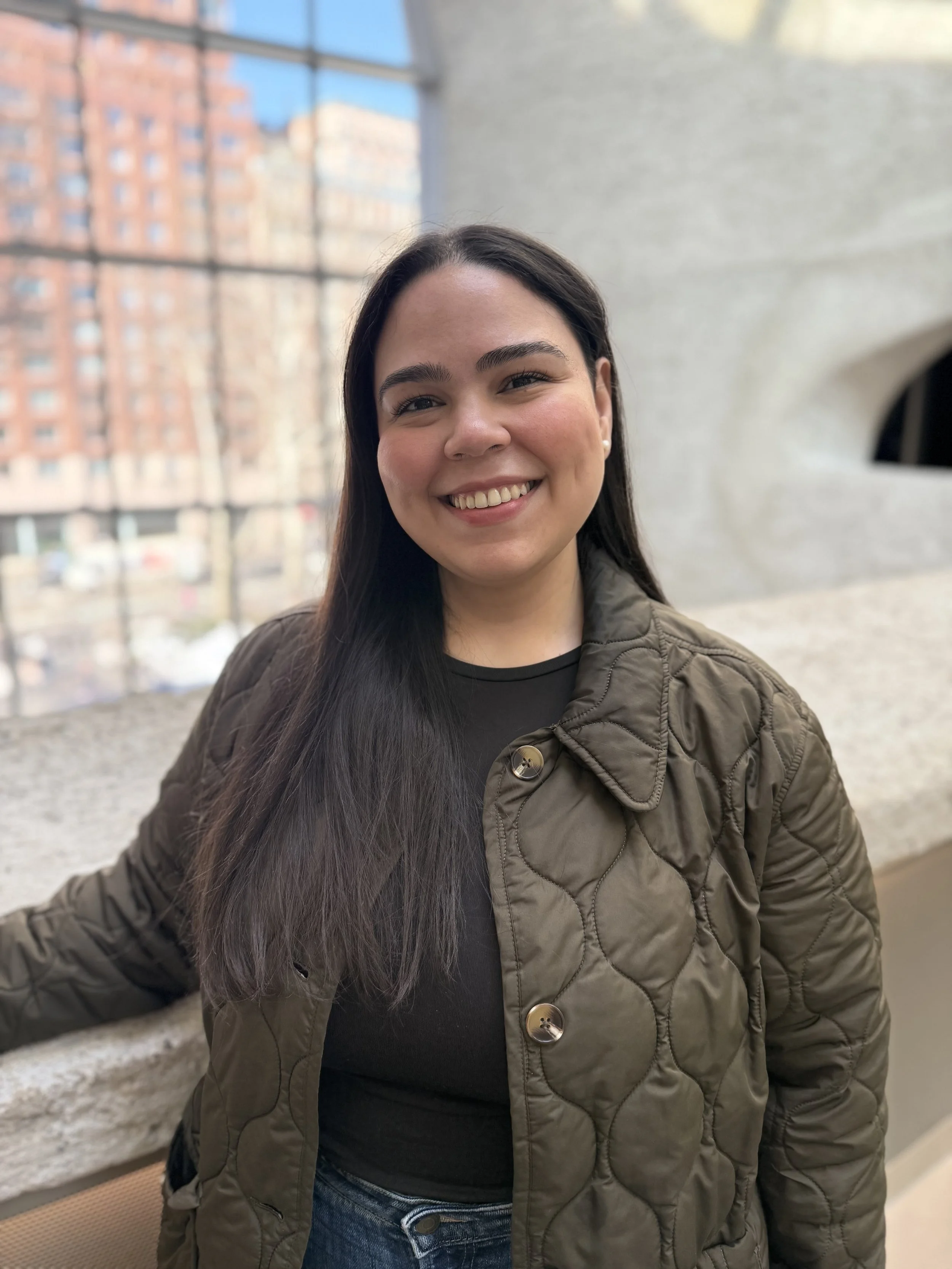 Smiling woman with long dark hair wearing an olive quilted jacket over a black top, standing indoors by a ledge with large windows and a city building blurred in the background.