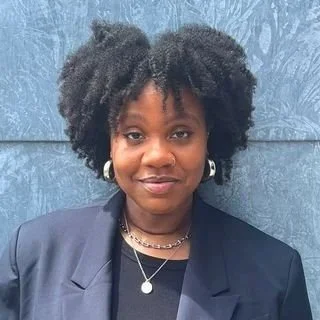 A smiling black woman with curly hair, wearing a dark blazer over a black top, silver hoop earrings and layered necklaces, standing in front of a textured blue wall.