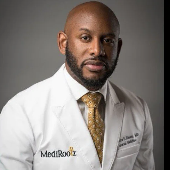 Black male physician with a closely trimmed beard wearing a white medical coat and patterned gold tie, posed against a neutral gray background.