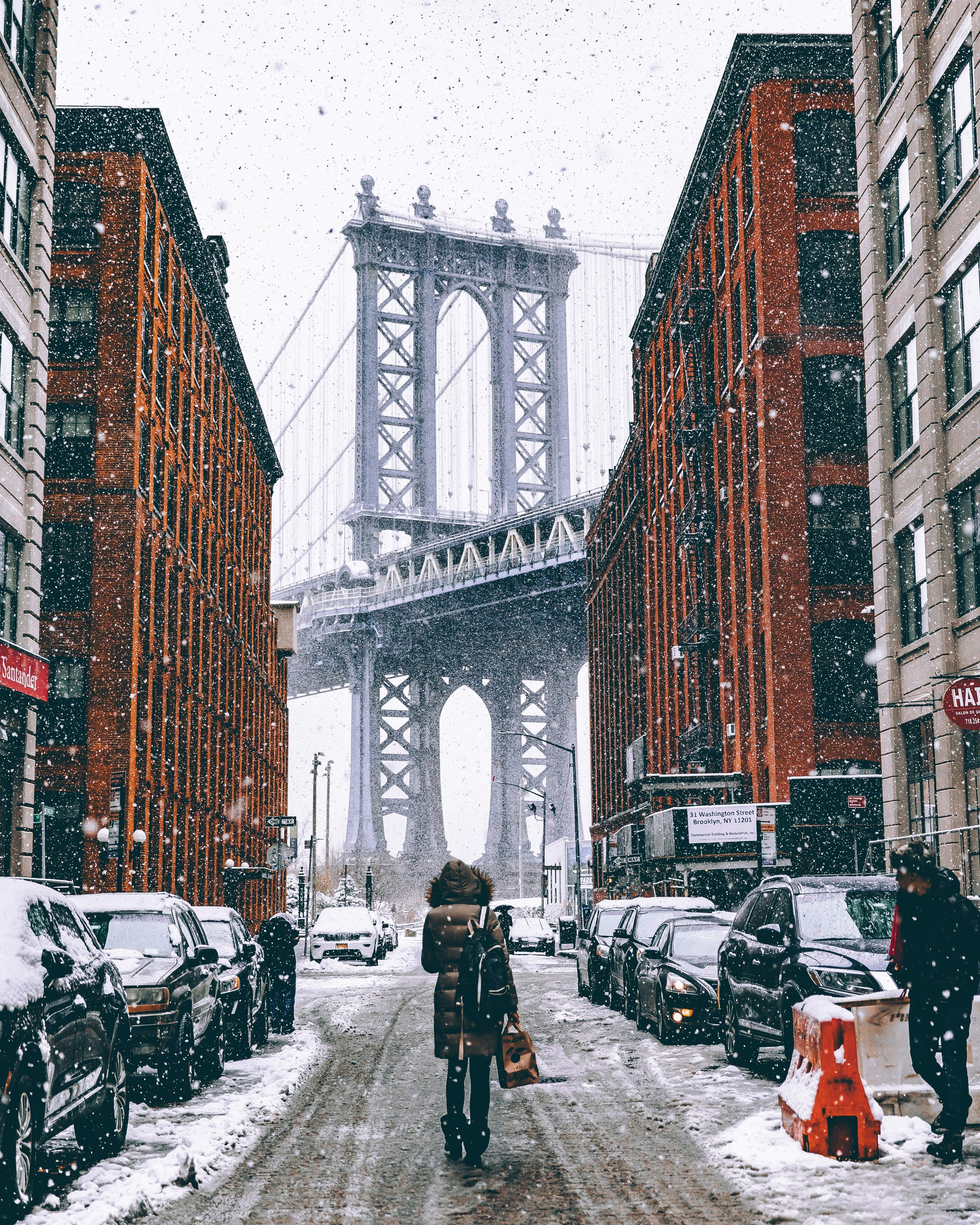A person walking alone down a snow-covered city street toward a large suspension bridge, framed by brick buildings and falling snow, evoking winter stillness, endurance, and movement through a cold, quiet moment.
